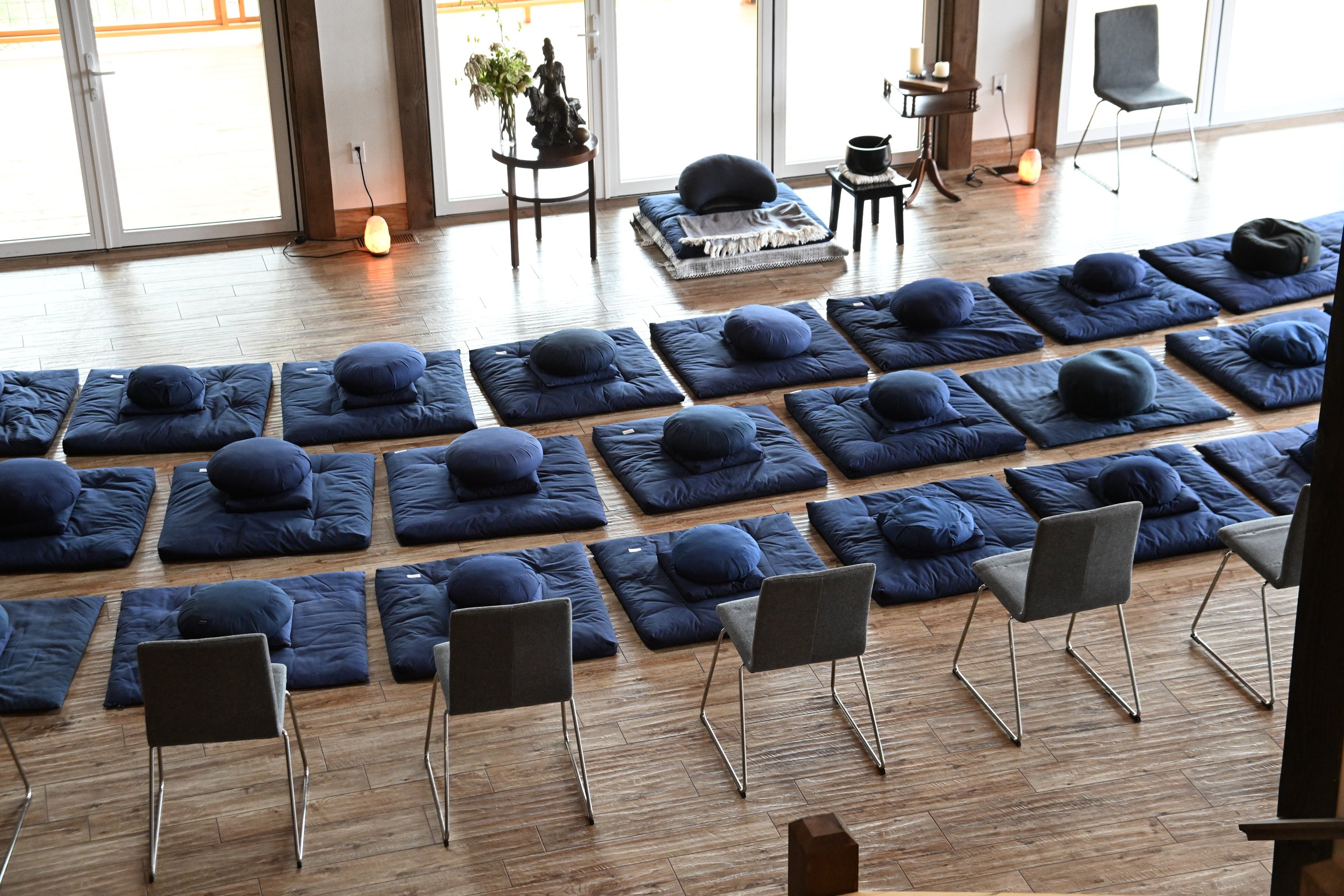 A room set up for a meditation or yoga session with blue cushions and round cushions arranged on a wooden floor, facing a sliding glass door. There are chairs around the room, and decorative items including candles, a chair with salt lamps, and plants are visible.
