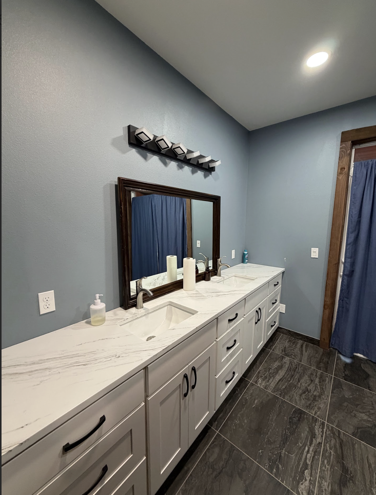 Bathroom with double vanity, marble countertop, framed mirror, blue walls, and dark floor tiles.