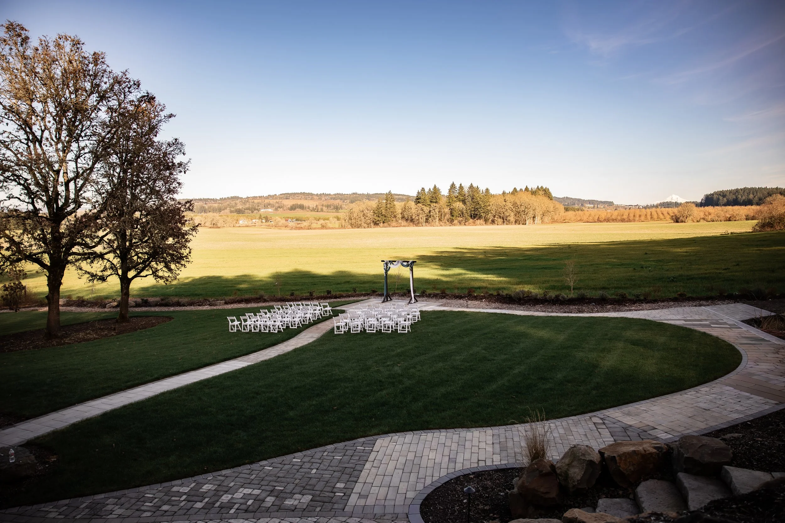 Outdoor wedding setup with white chairs arranged in rows on green grass, a small arch, and a scenic view of rolling hills and trees in the background on a clear day.