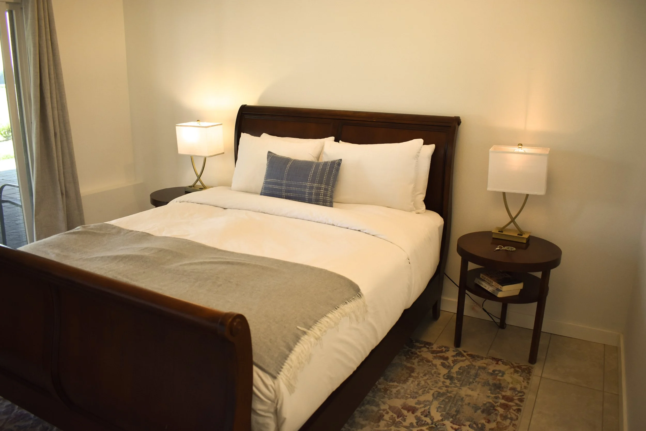 Hotel bedroom with a large bed, white pillows, a blue decorative pillow, bedside lamps, a nightstand, and a patterned area rug.