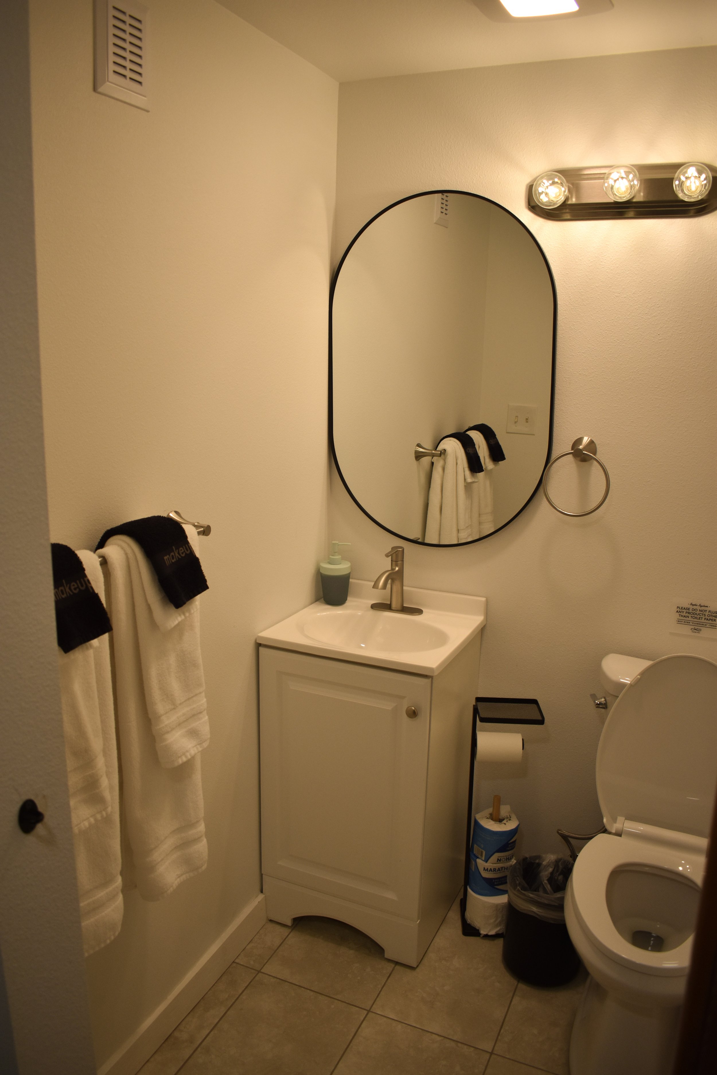 A small bathroom with a white vanity sink, an oval mirror, a wall-mounted light fixture with four bulbs, a towel ring with a towel, a toilet with an open lid, and various toiletries and waste bins. The walls are painted light beige, and the floor is tiled in neutral tones.