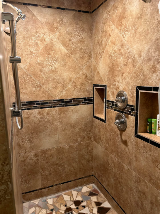 Bathroom shower with beige tiled walls, a decorative black tile border, two recessed shelves holding toiletries, and a geometric patterned tile floor.