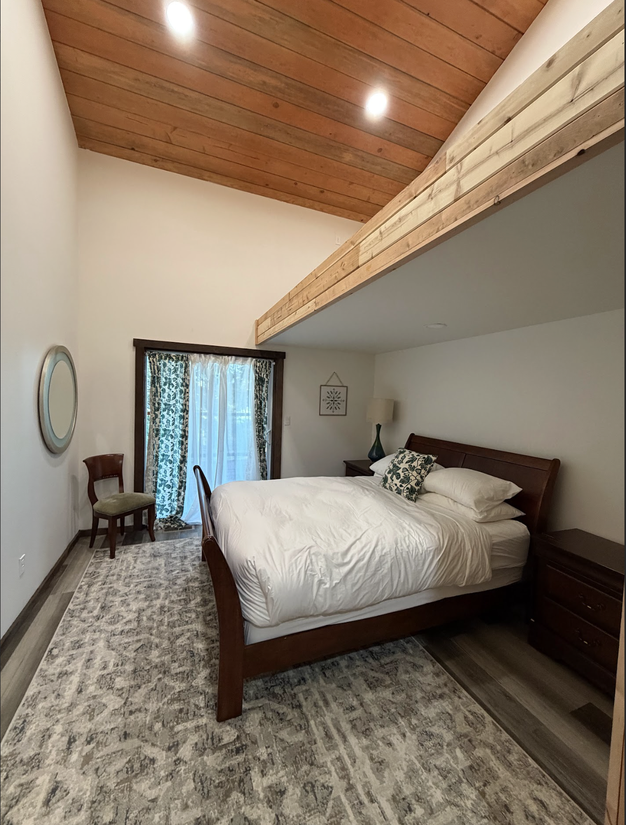 A bedroom with a wooden ceiling, a bed with white bedding, a nightstand with a lamp, a window with curtains, an armchair, and patterned carpet.