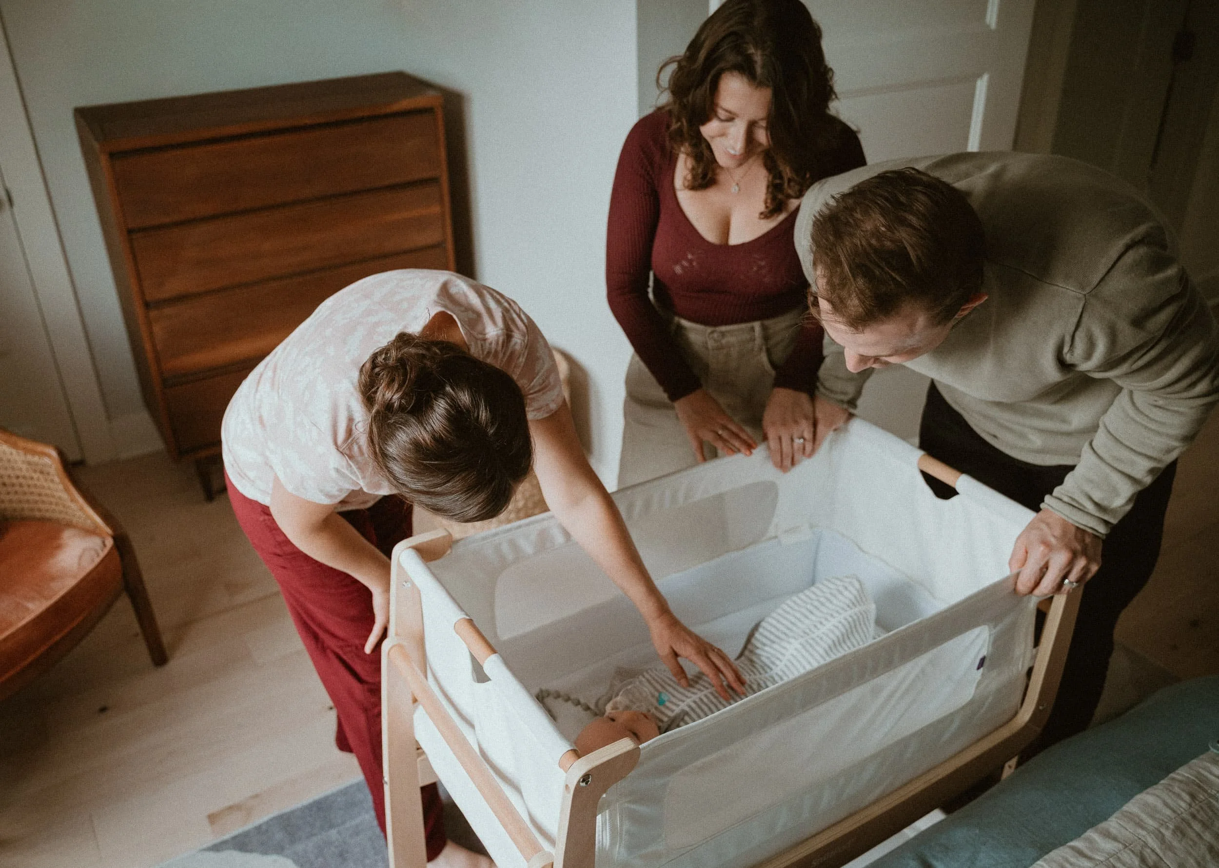 Julia-Rose Scianna discusses sleep consulting services with new parents in Portland while watching over a baby in a crib.