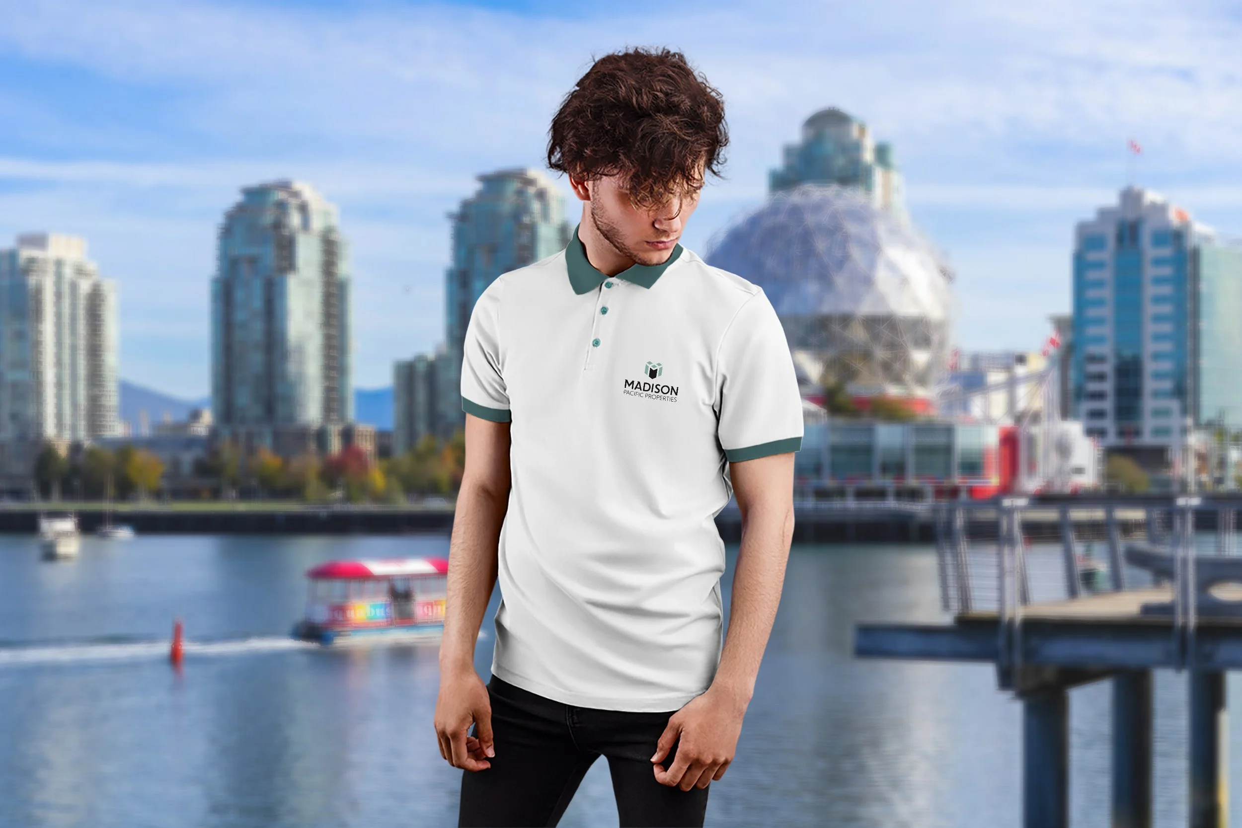 Young man with curly hair in a white polo shirt with green collar and sleeve cuffs, branded 'Madison Pacific Properties', standing outdoors in front of a city skyline and water, with boats and a ride visible in the background.