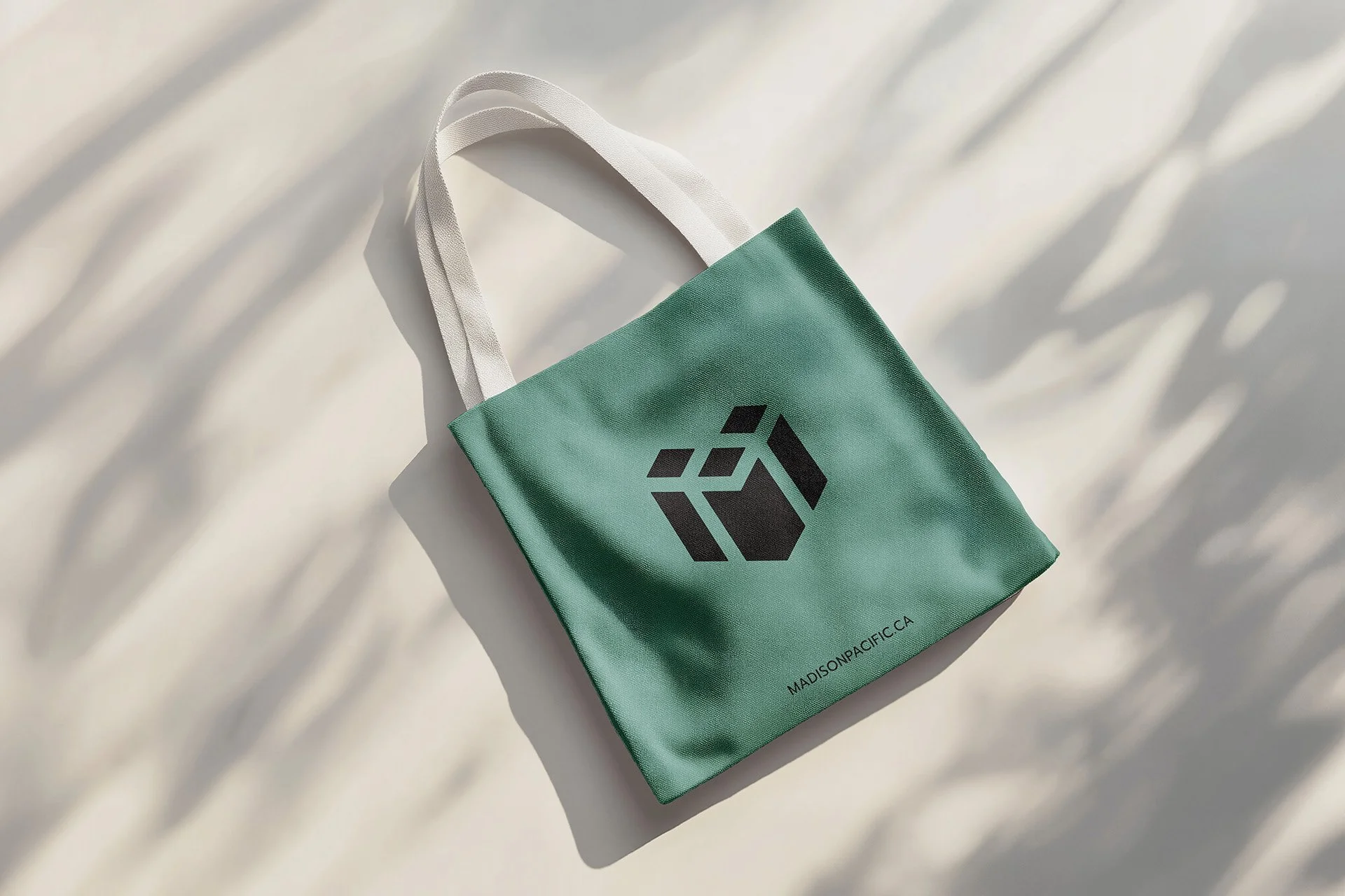A teal reusable shopping bag with a black geometric logo on the front, lying on a white surface. The bag's handles are made of white fabric and cast shadows on the surface. The bottom edge of the bag has text that reads "MADISONPACIFIC.CA."