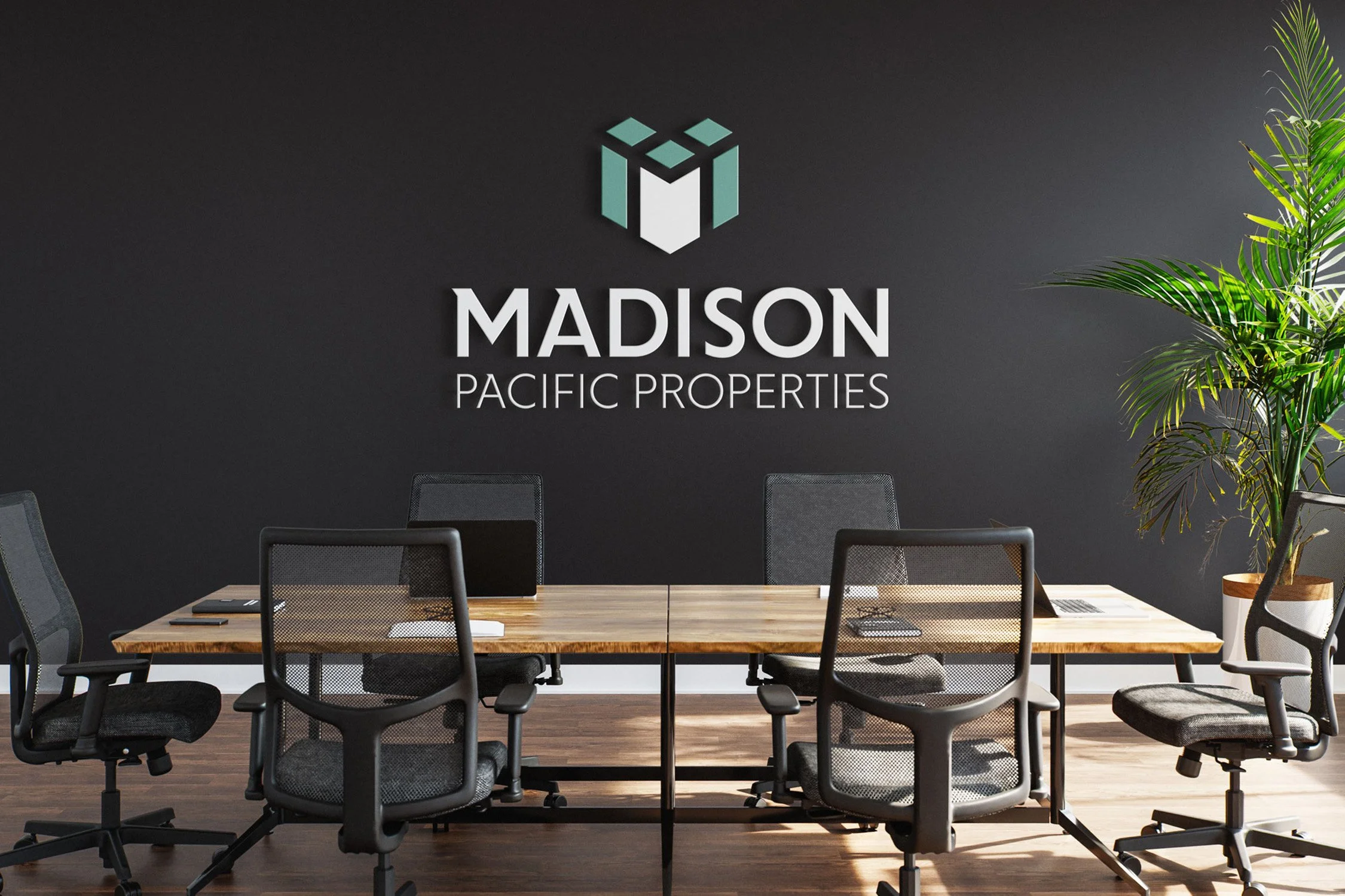 Conference room with a wooden table, black office chairs, a wall with the logo 'Madison Pacific Properties,' a large potted plant, and a dark wall.