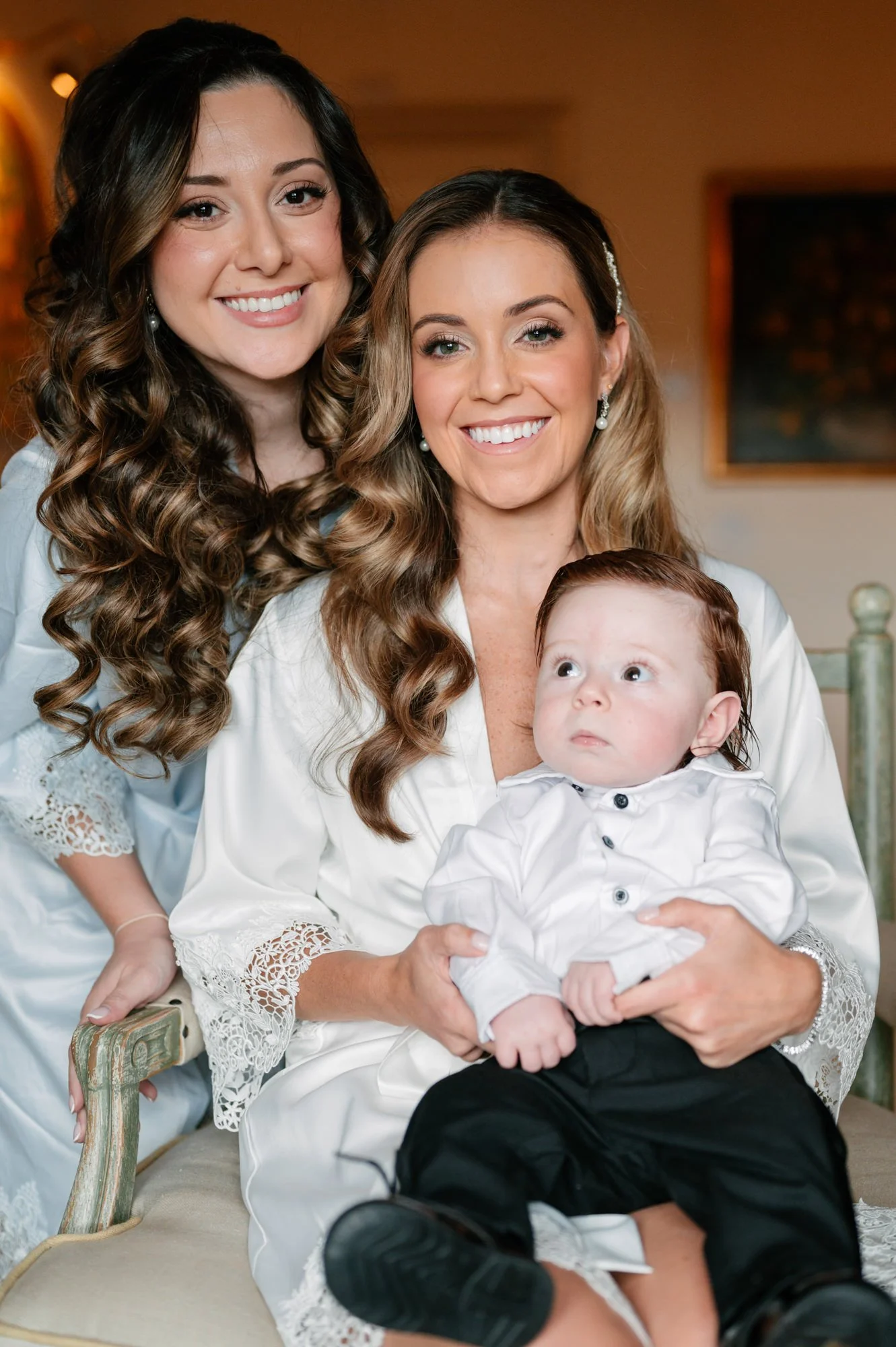 Three women, two with long curly hair and one with short brown hair, sitting together. The woman with short hair is holding a young boy with red hair dressed in a white shirt and black pants. All are smiling, with the boy looking up.