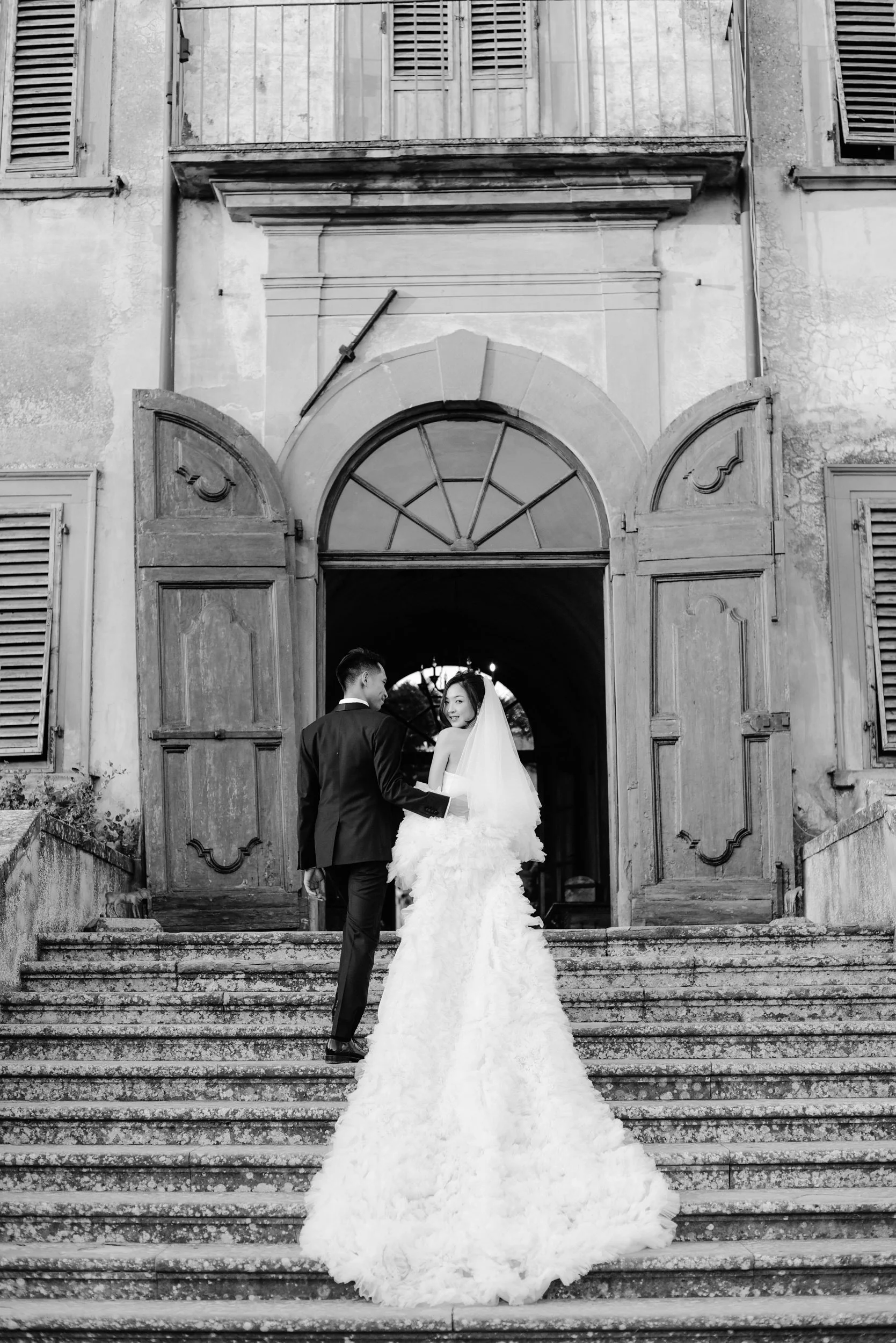 Bride and groom on outdoor steps in front of old building with open doors, bride in wedding gown with veil, groom in tuxedo, black and white photo.