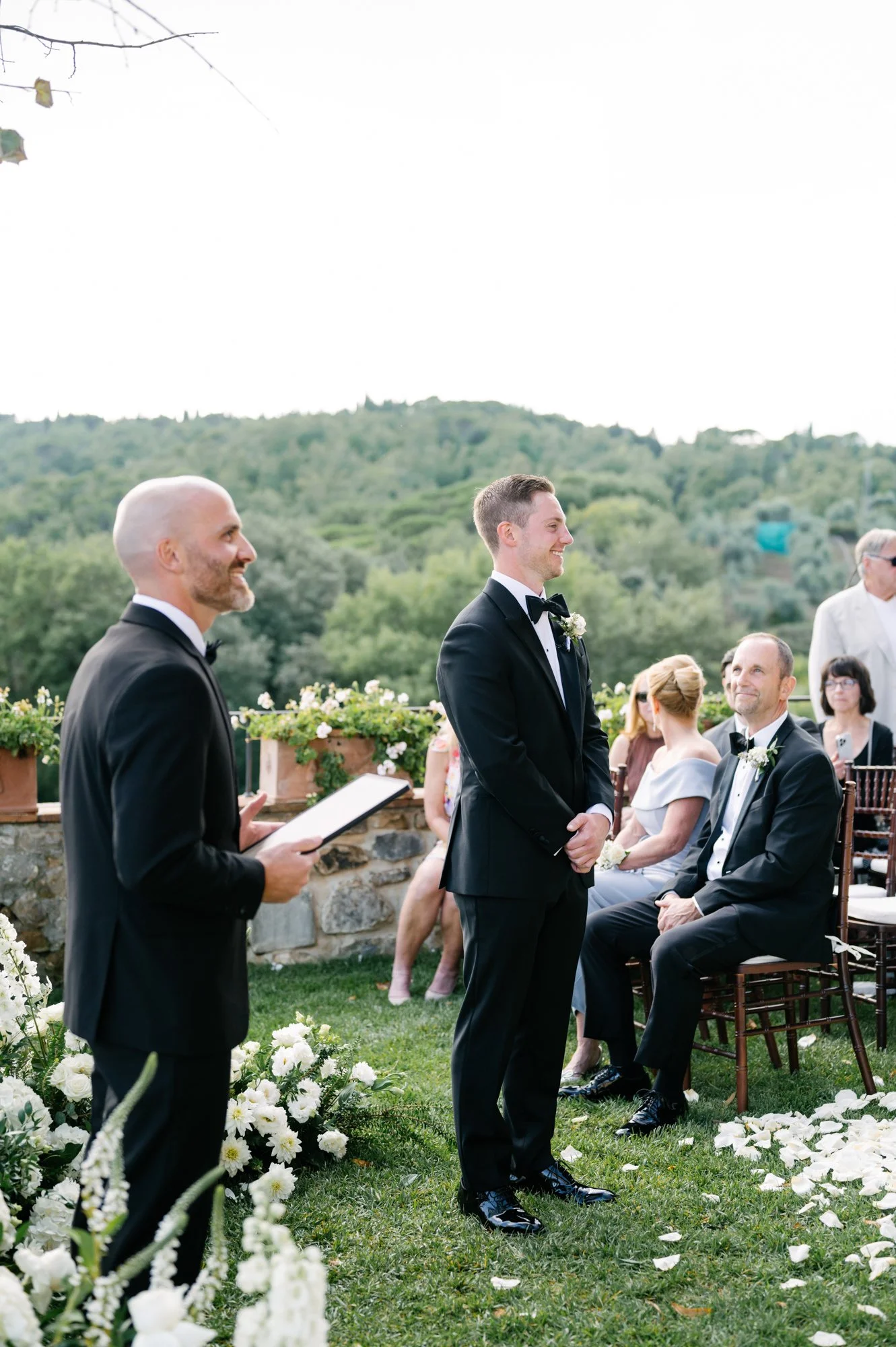 A wedding ceremony outdoors with a groom standing in front of an officiant, surrounded by guests, flowers, and greenery in the background.