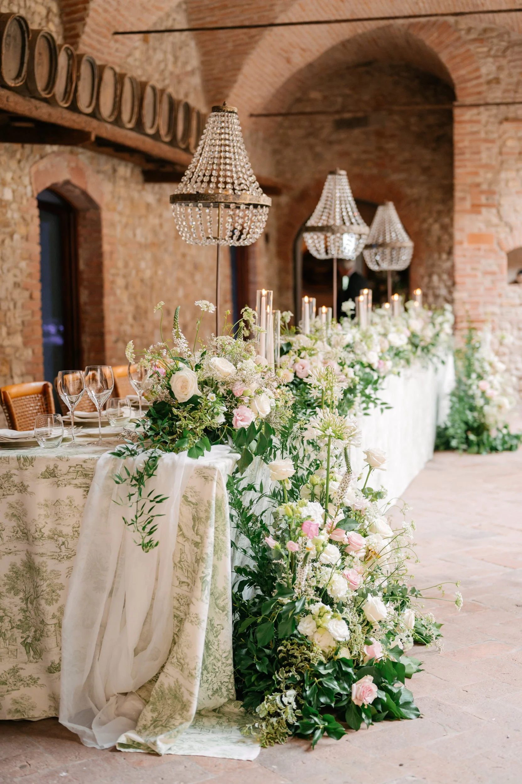 Elegant wedding or event table decorated with white and pink flowers, candles, and crystal chandeliers in a rustic brick venue.