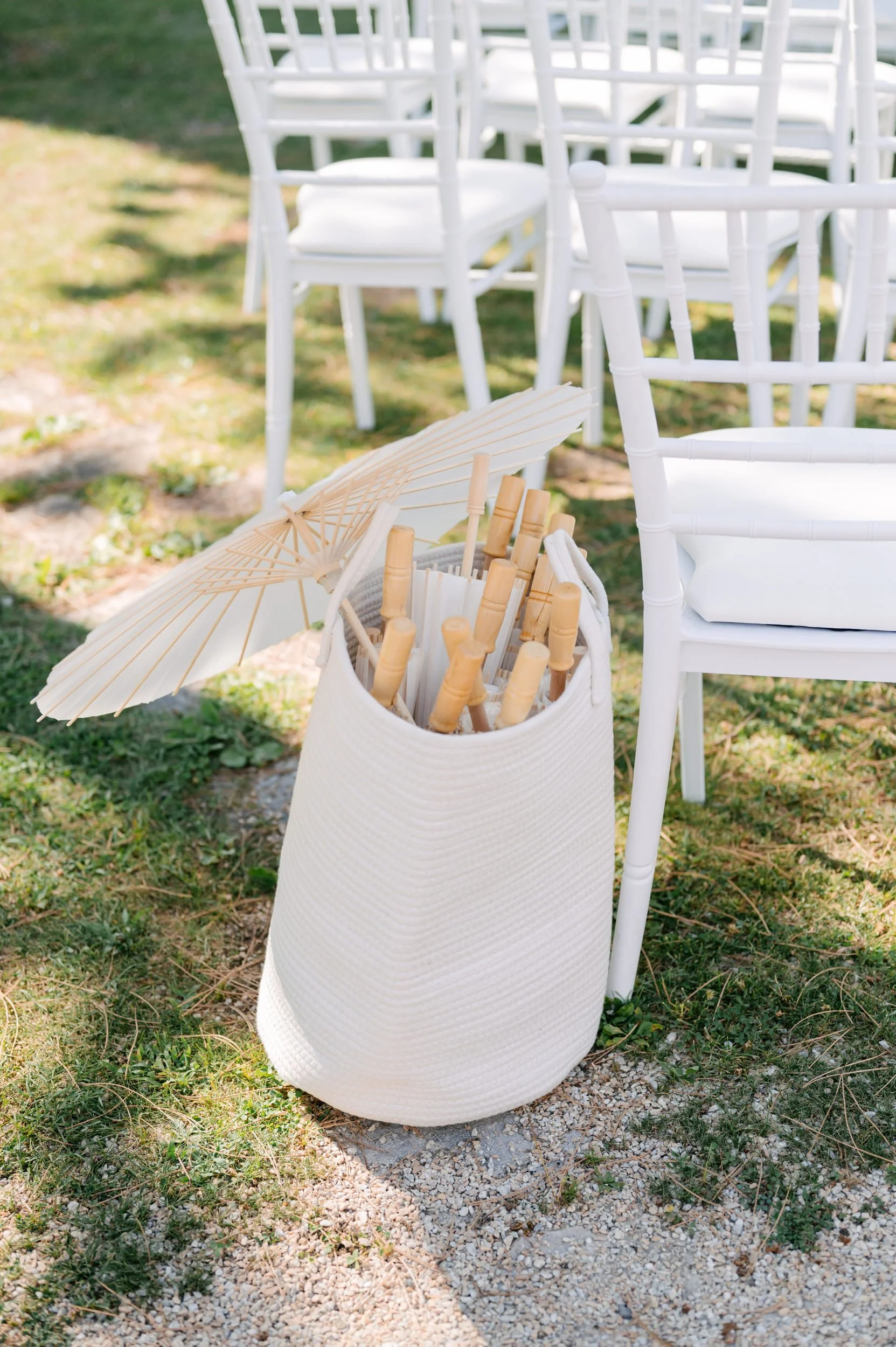 Empty white chair setup outdoors with a large white basket filled with paper umbrellas and bamboo fans beside it.