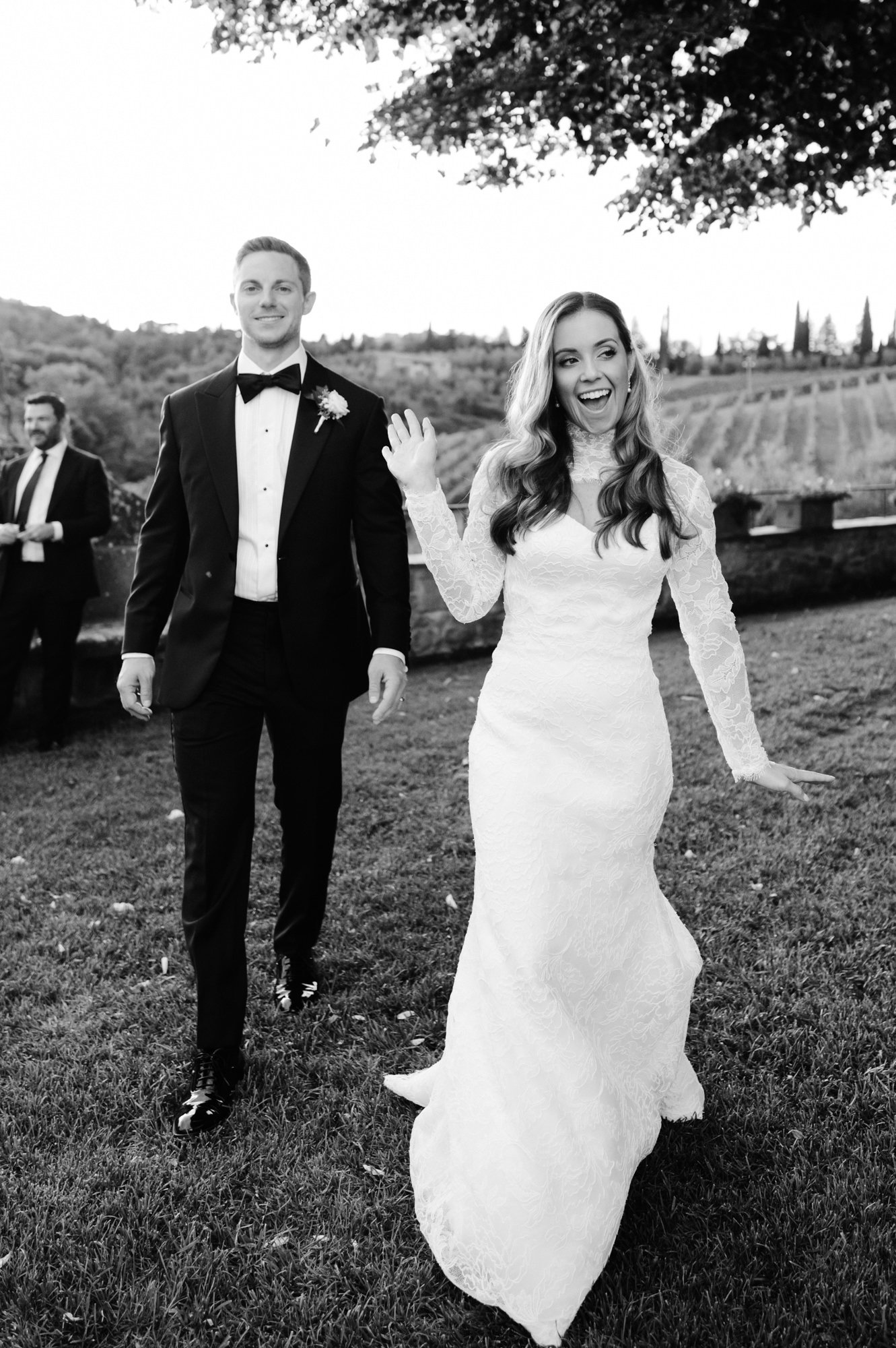 Black and white photo of a bride in a white lace wedding dress smiling and waving, walked outdoors on grass, with a groom in a tuxedo walking behind her and other people in the background, scenic landscape with trees and hills.