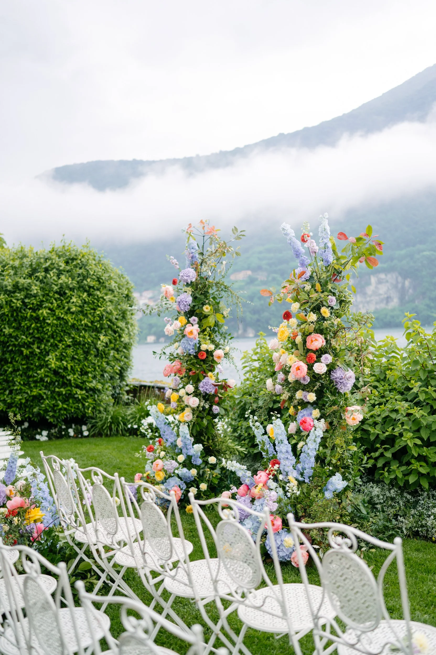 Wedding ceremony setup with white chairs facing floral arch and scenic mountain view with lake and clouds in the background.