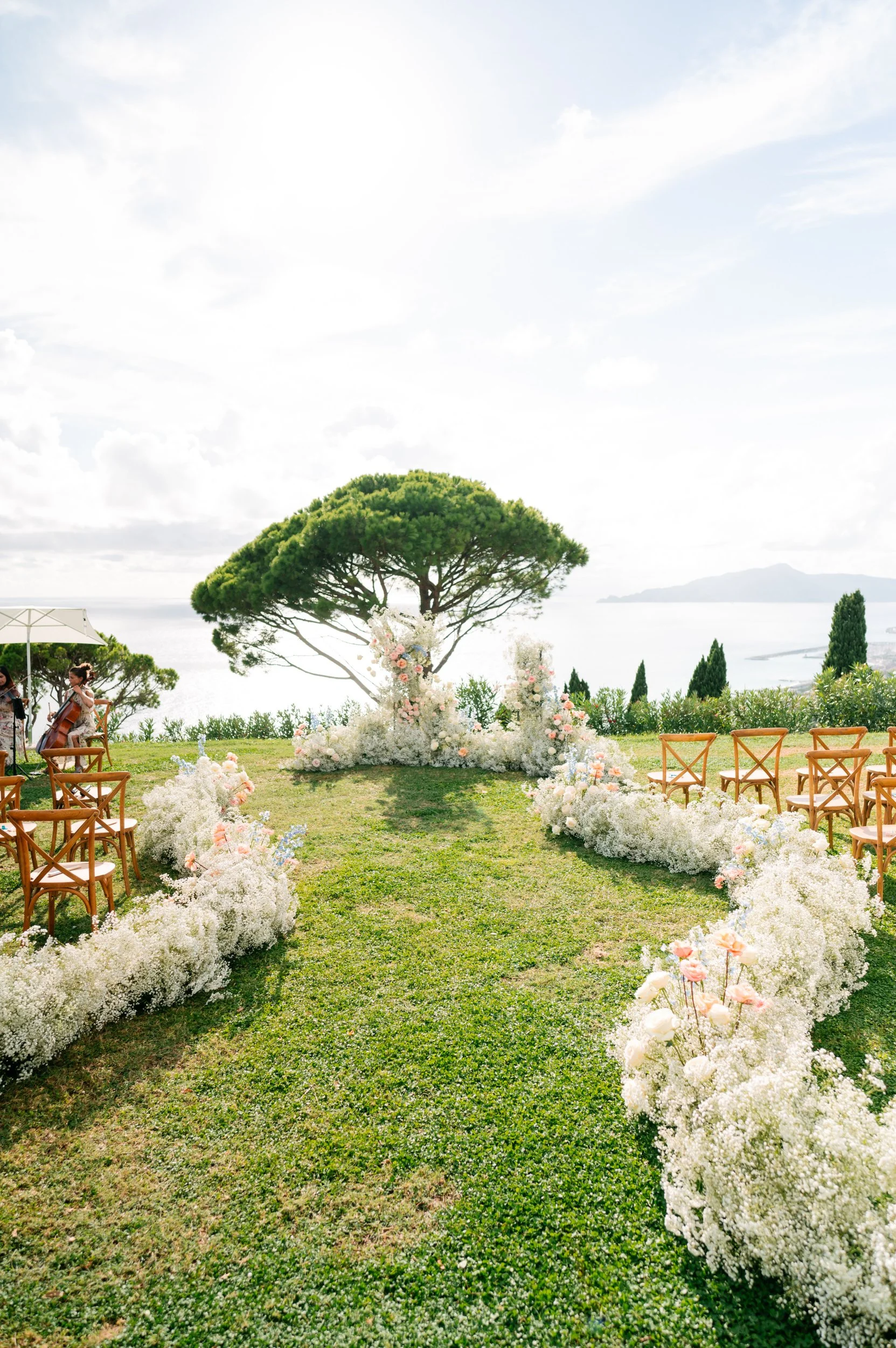 Outdoor wedding ceremony setup with rows of wooden chairs, floral arrangements lining the aisle, and a large tree at the altar on a grassy hill overlooking water and mountains in the distance.