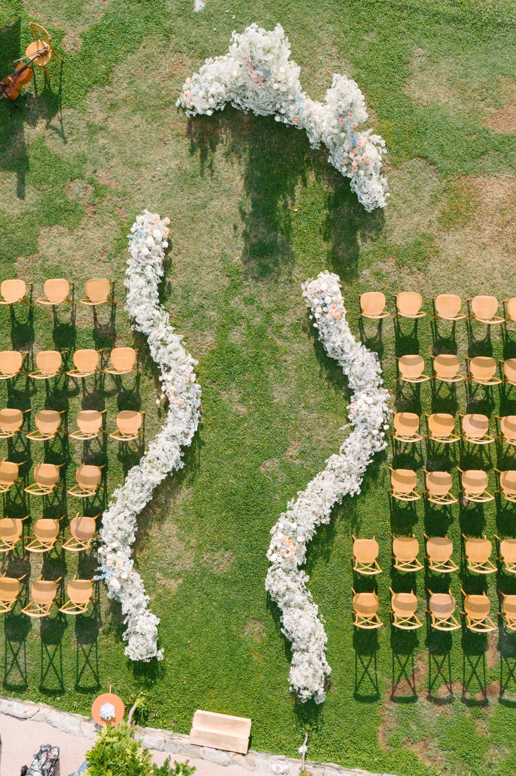 Aerial view of an outdoor wedding setup with rows of tan chairs on green grass, and a flower arch shaped like a silhouette of a woman's profile.