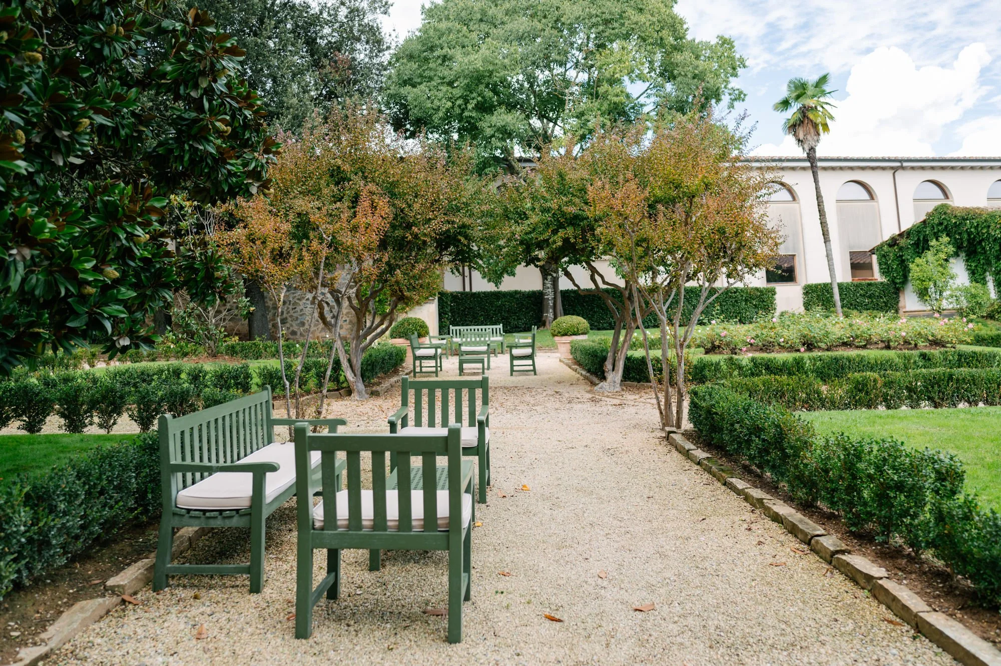 Garden with green benches and trees along a gravel pathway, with manicured bushes and a white building in the background.