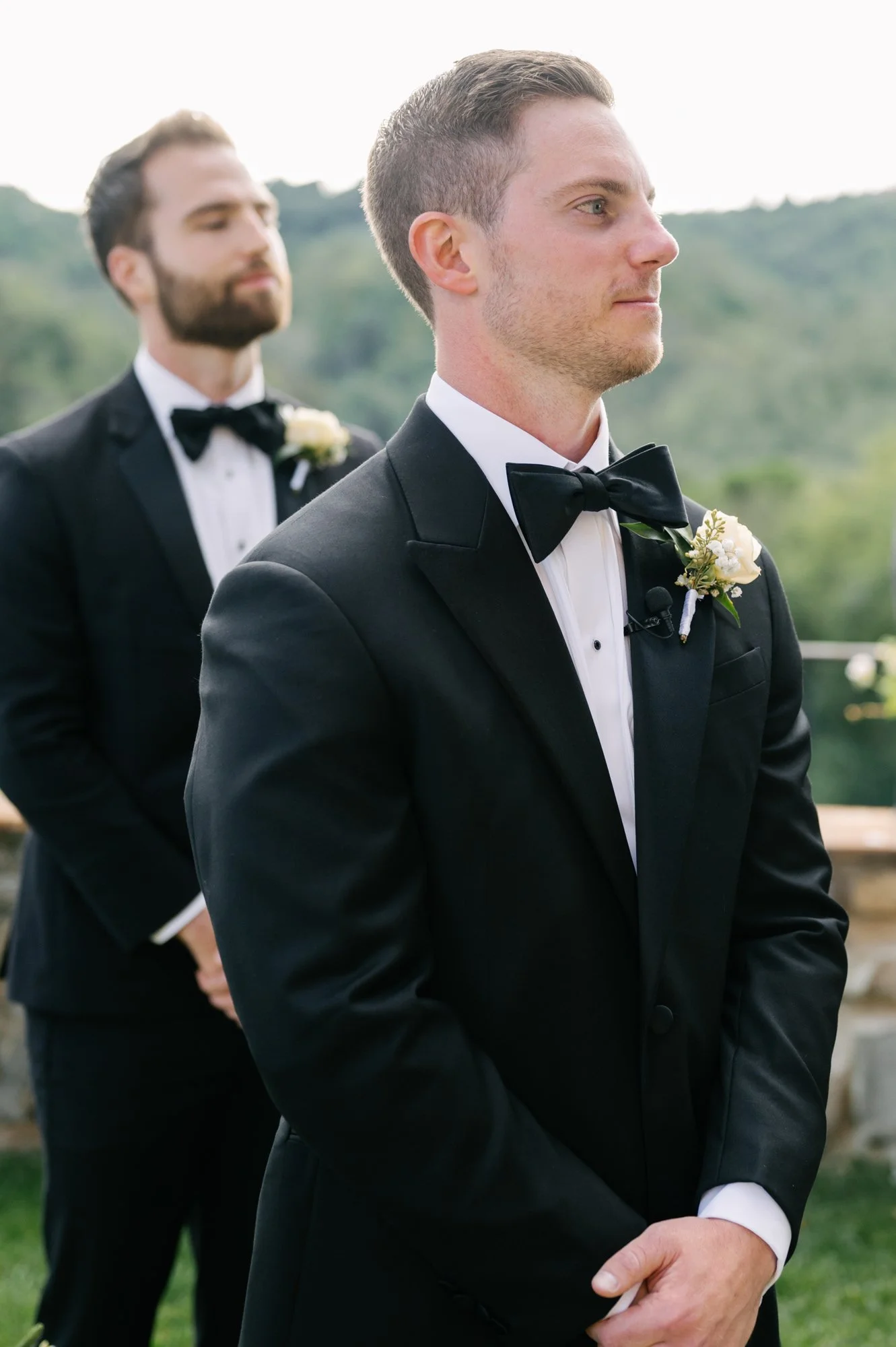 Two men in black tuxedos and bow ties standing outdoors with a scenic background; one is in front, the other slightly behind and blurred.