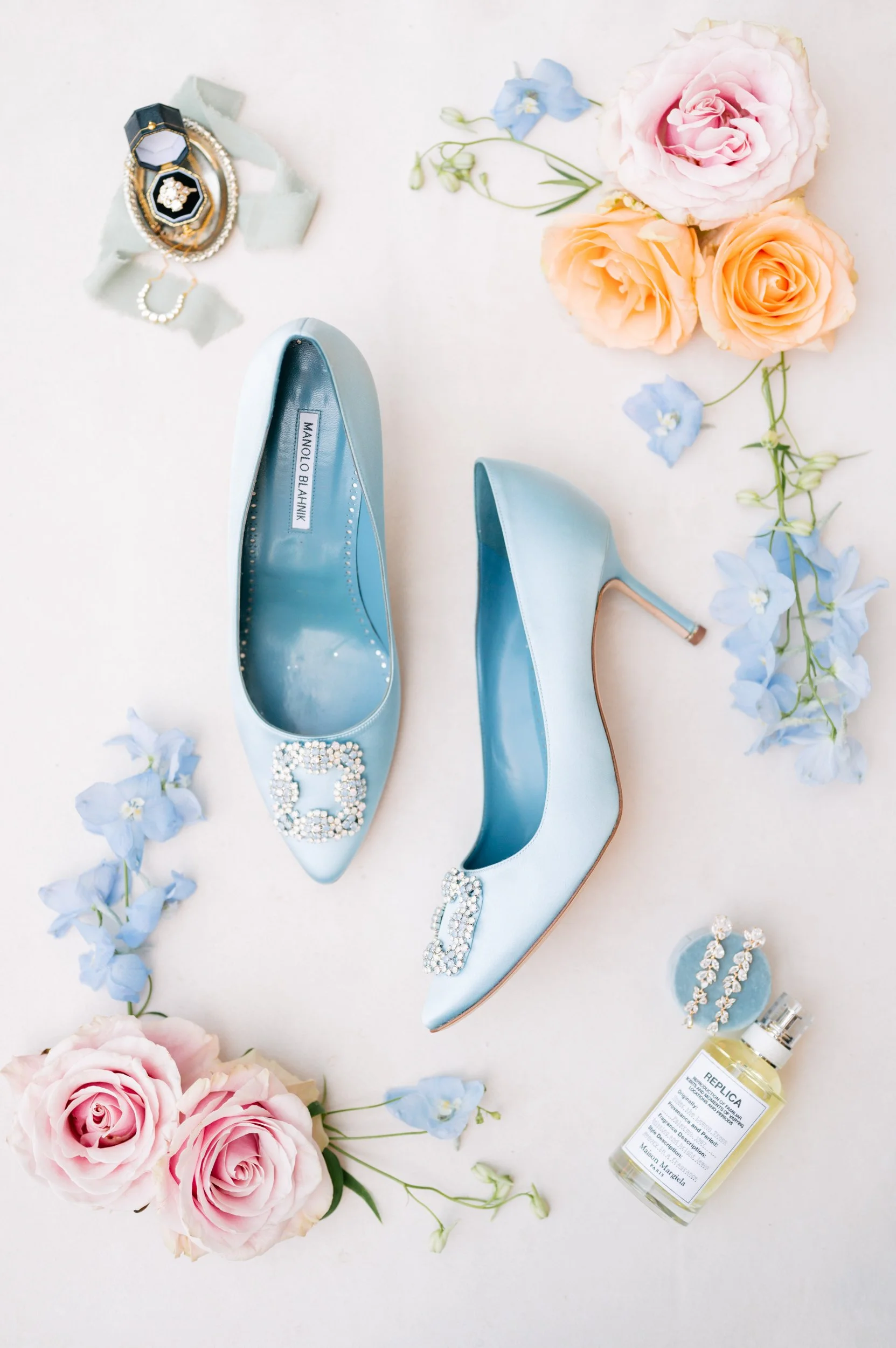 Light blue designer high heel shoes with rhinestone embellishments, surrounded by pink roses, blue flowers, jewelry, and a perfume bottle on a white background.