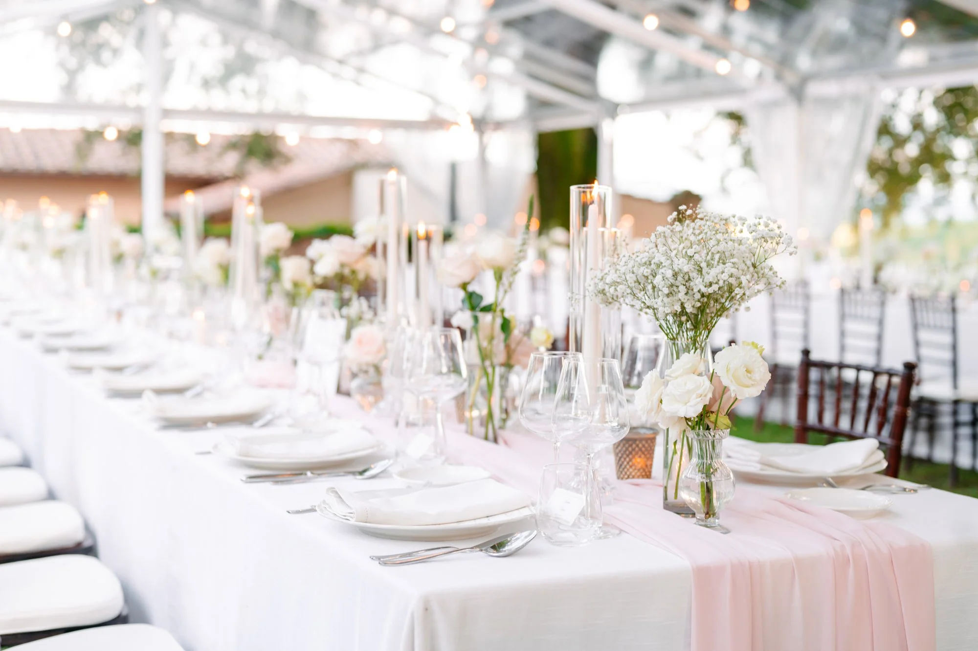 Elegant outdoor event table setup with white tablecloth, floral centerpieces, tall candles, glassware, and place settings, under a tent with string lights. Luxury editorial wedding photography in Tuscany. Destination wedding in Italy. L&V Photography
