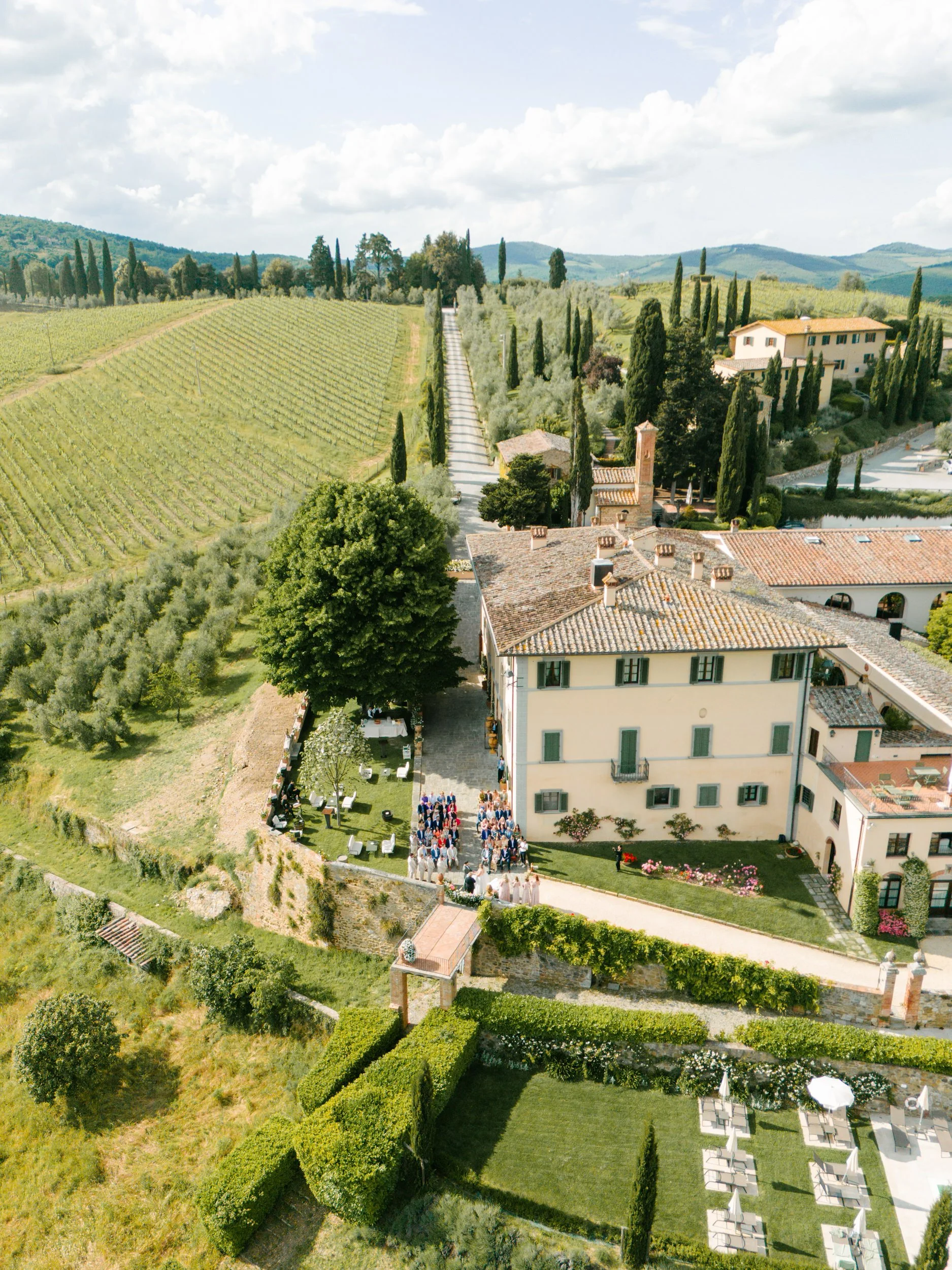 An aerial view of a large house with people gathering outside on a sunny day. Surrounding the house are lush green gardens, trees, and fields with rows of crops, all set in a scenic countryside with rolling hills in the background.