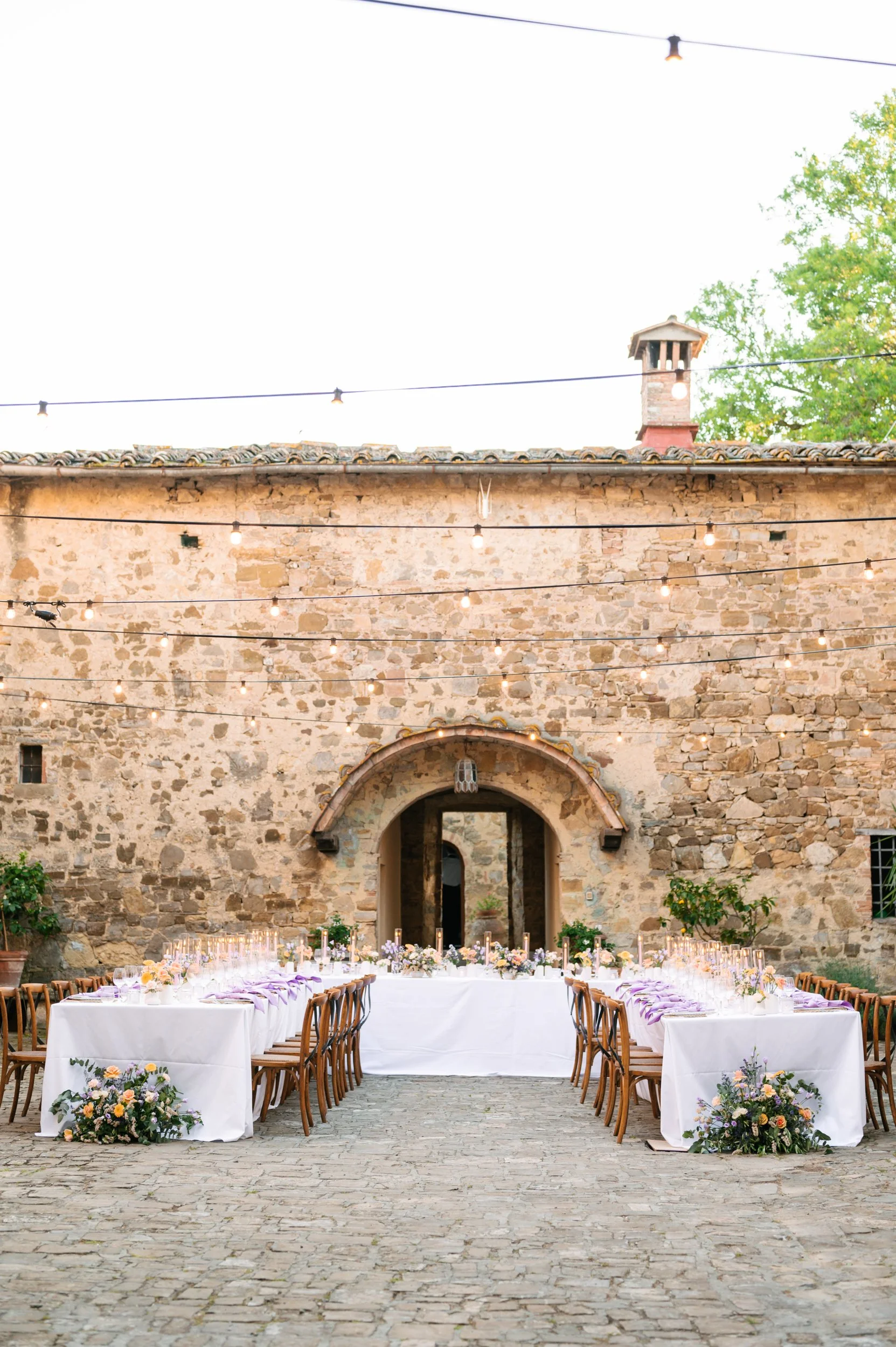 Outdoor wedding reception setup with long tables draped in white cloth, floral arrangements, and candles, set against a rustic stone wall decorated with string lights at dusk.
