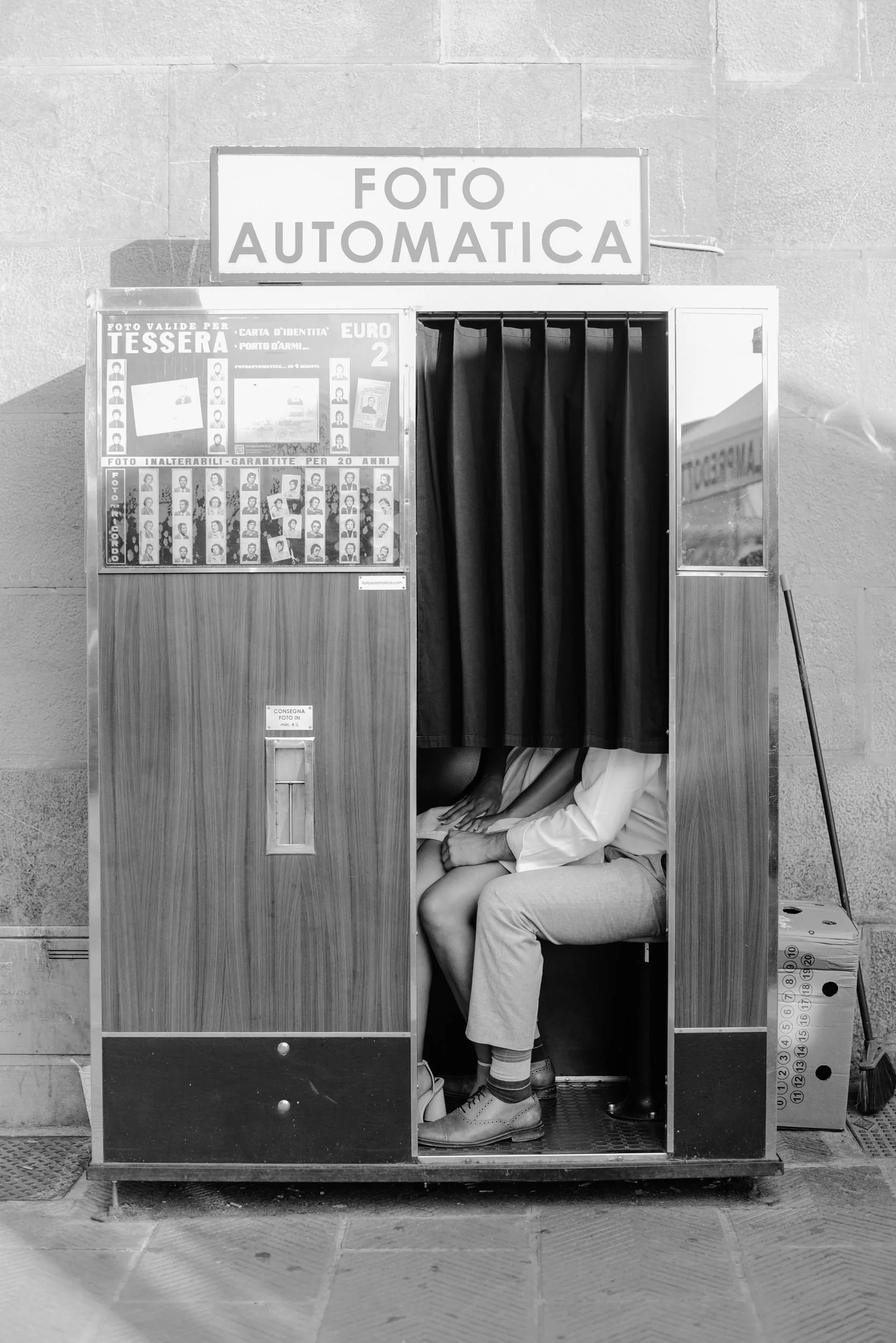 Black and white photo of a photo booth with two people inside, one sitting and the other leaning over. The booth has a sign on top reading 'FOTO AUTOMATICA' and a poster on the left side with headshots of various people.