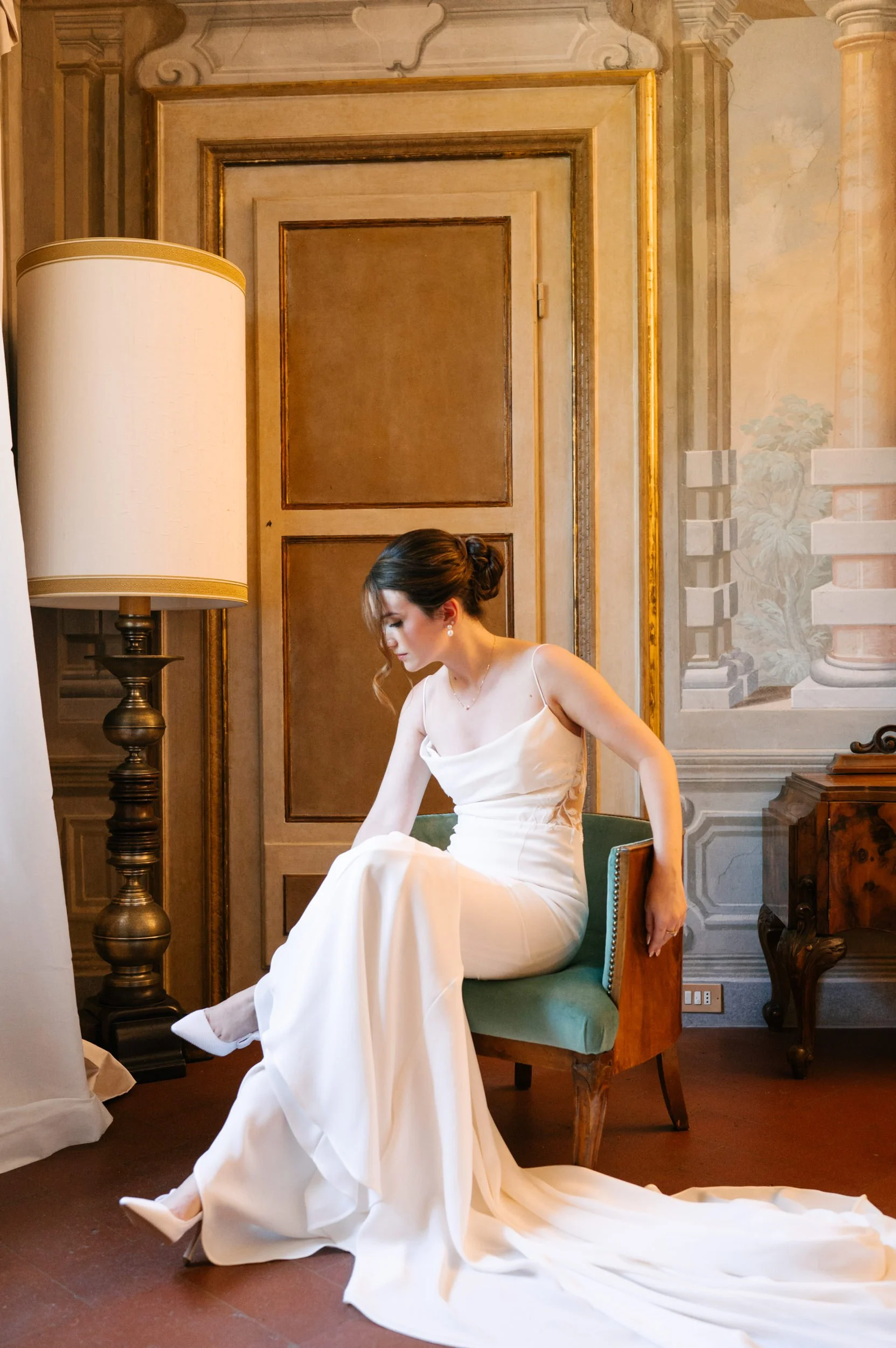 A woman in a white dress, sitting on a green cushioned chair, adjusting her shoe in an ornate room with wood paneling, gold accents, and classical wall paintings.