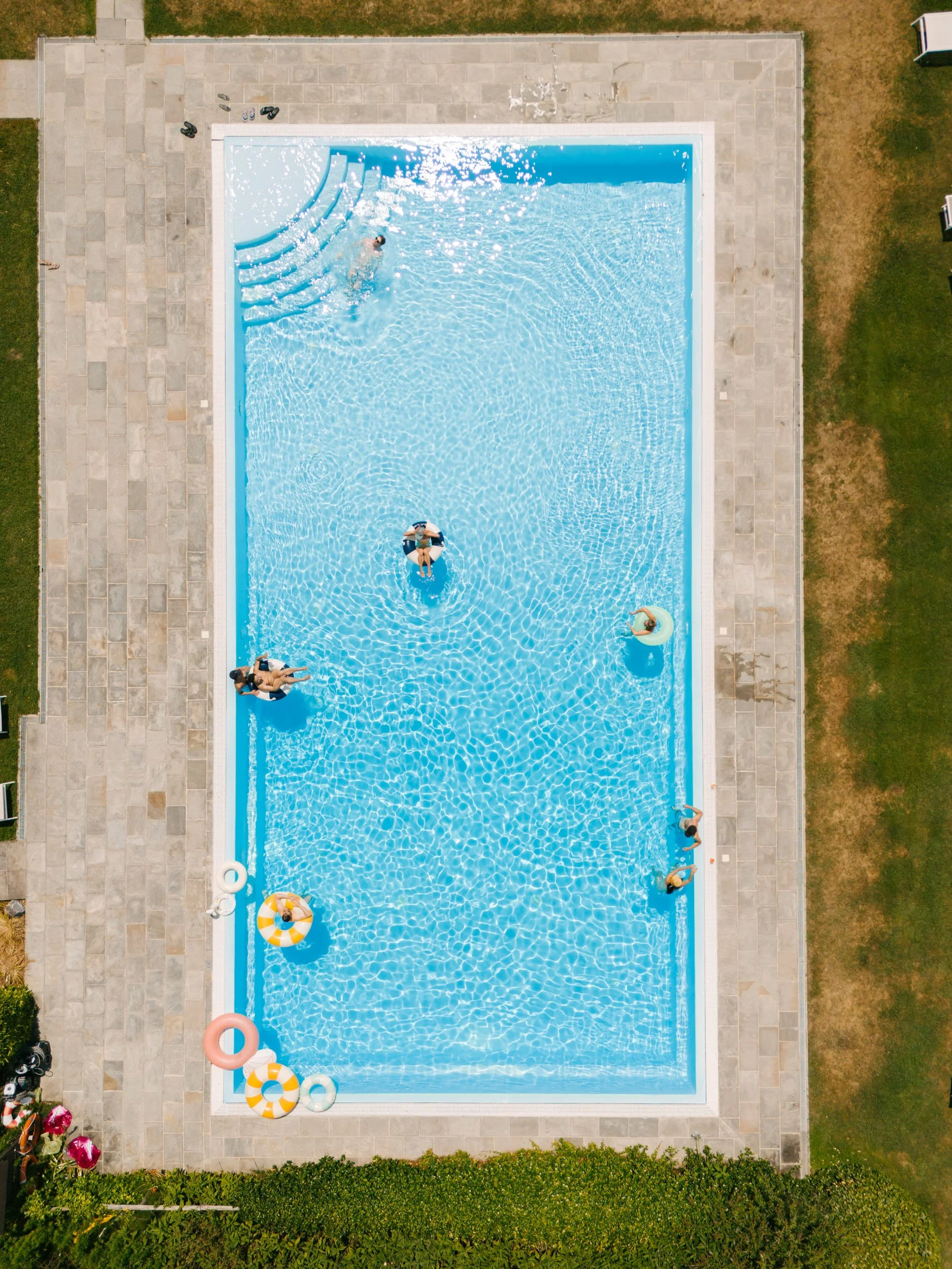 Aerial view of a rectangular outdoor swimming pool with people swimming and floating, surrounded by a stone deck and grassy areas.