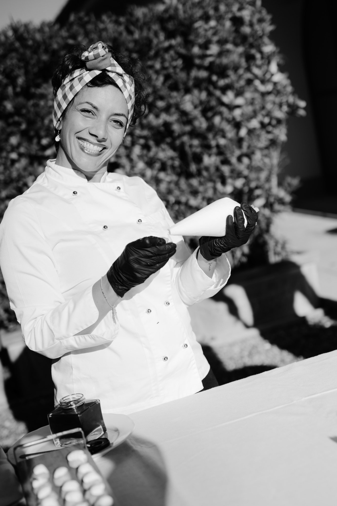 A woman wearing a chef's coat, black gloves, and a checkered headband is smiling and holding a piping bag, with a Christmas tree in the background.