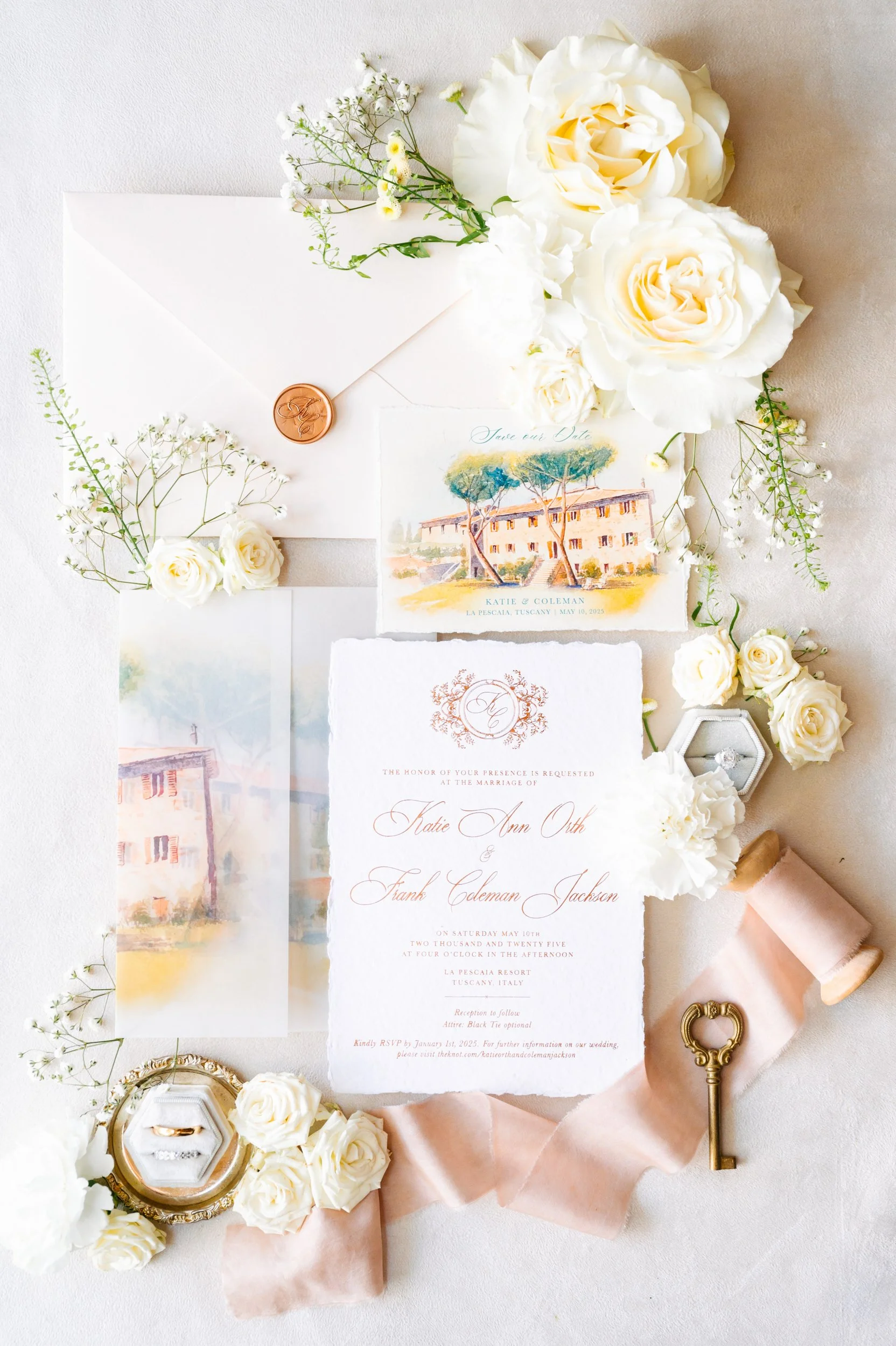 Wedding invitation surrounded by white roses, baby's breath, a wrapped pink ribbon, a vintage key, wedding rings in a jewelry box, and a small watercolor painting of a Tuscan villa.