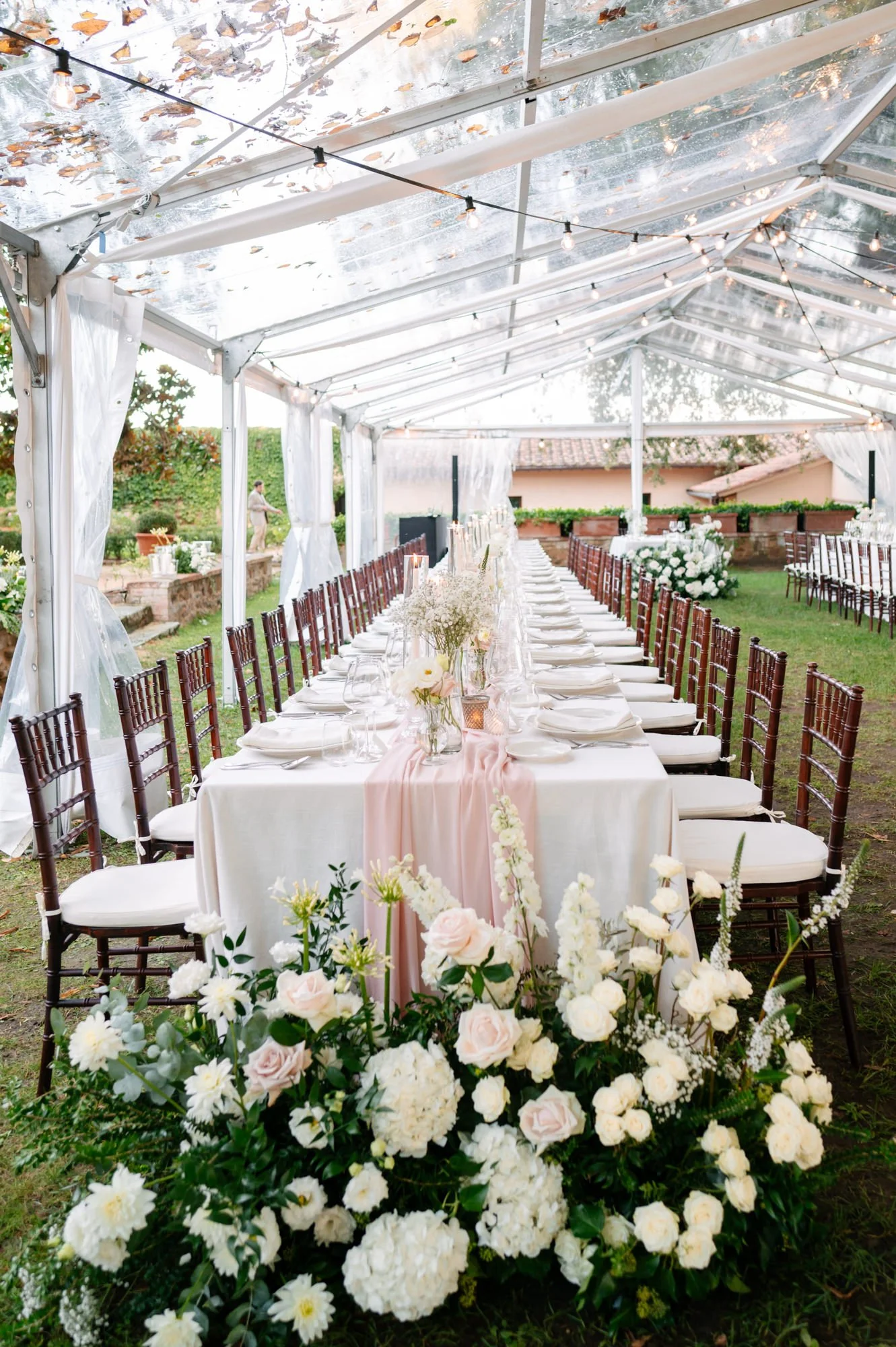 Elegant outdoor wedding reception under a transparent tent with string lights, a long rectangular table with white tablecloth, pink runner, floral centerpieces, and surrounded by wooden chairs, decorated with white and blush flowers.
