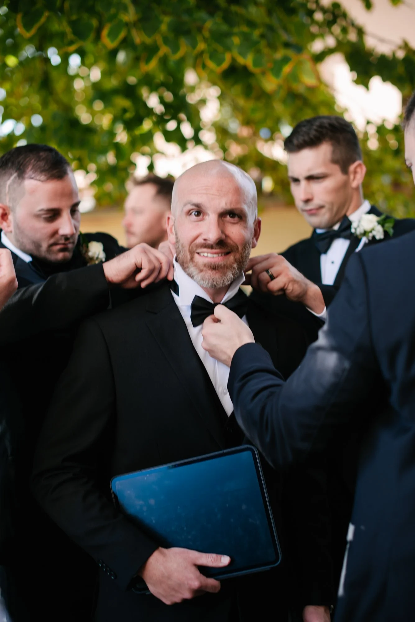 Man in a tuxedo with a bow tie being assisted by other men in tuxedos, one holding a tablet, as they prepare him for a formal event, possibly a wedding.