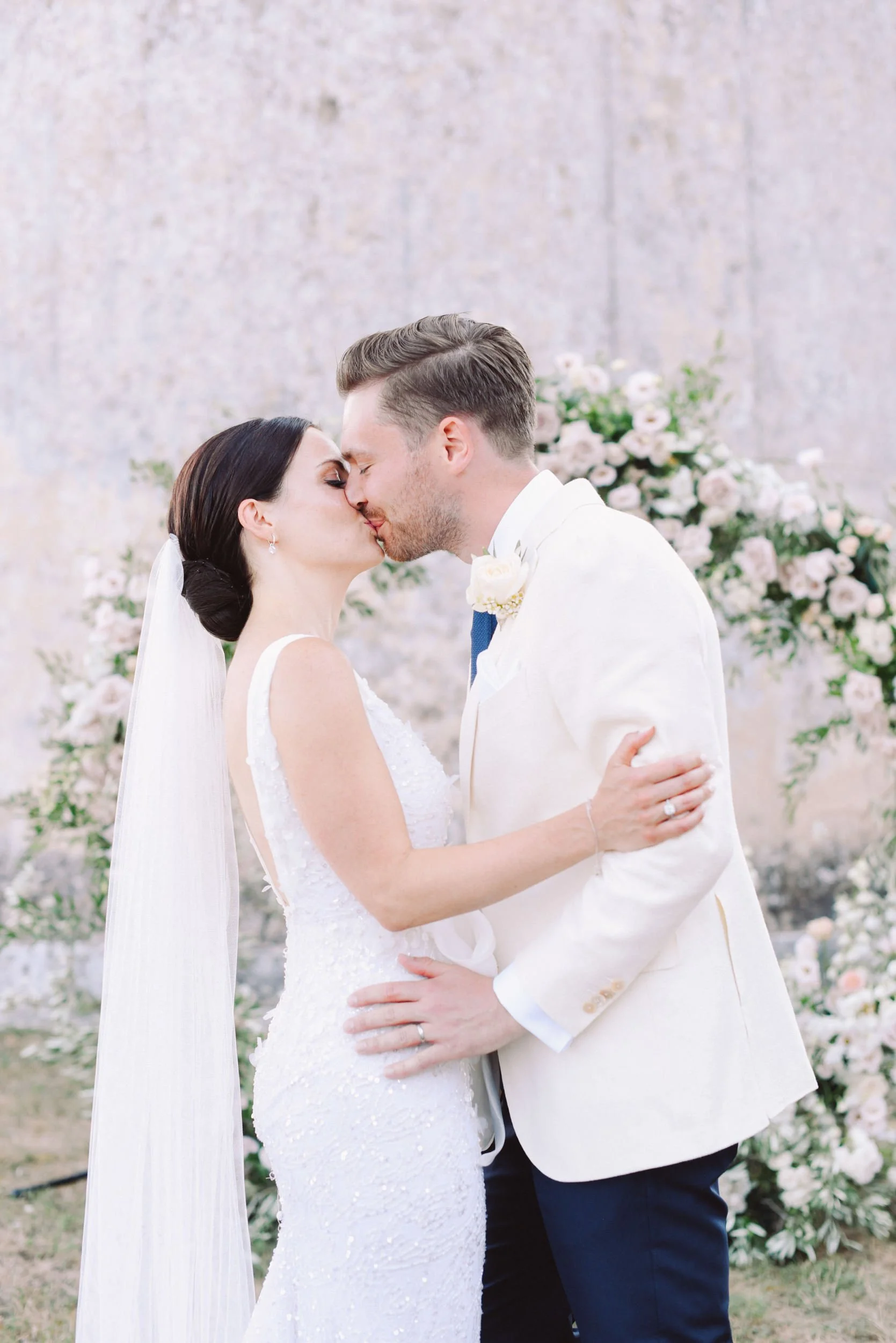 A bride and groom kiss at their wedding, with floral arrangements in the background.