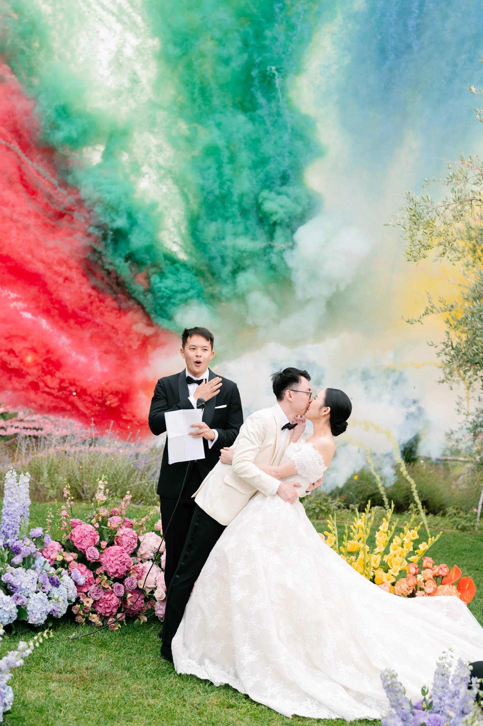 Wedding couple kissing with a colorful smoke display in the background, surrounded by vibrant flowers.