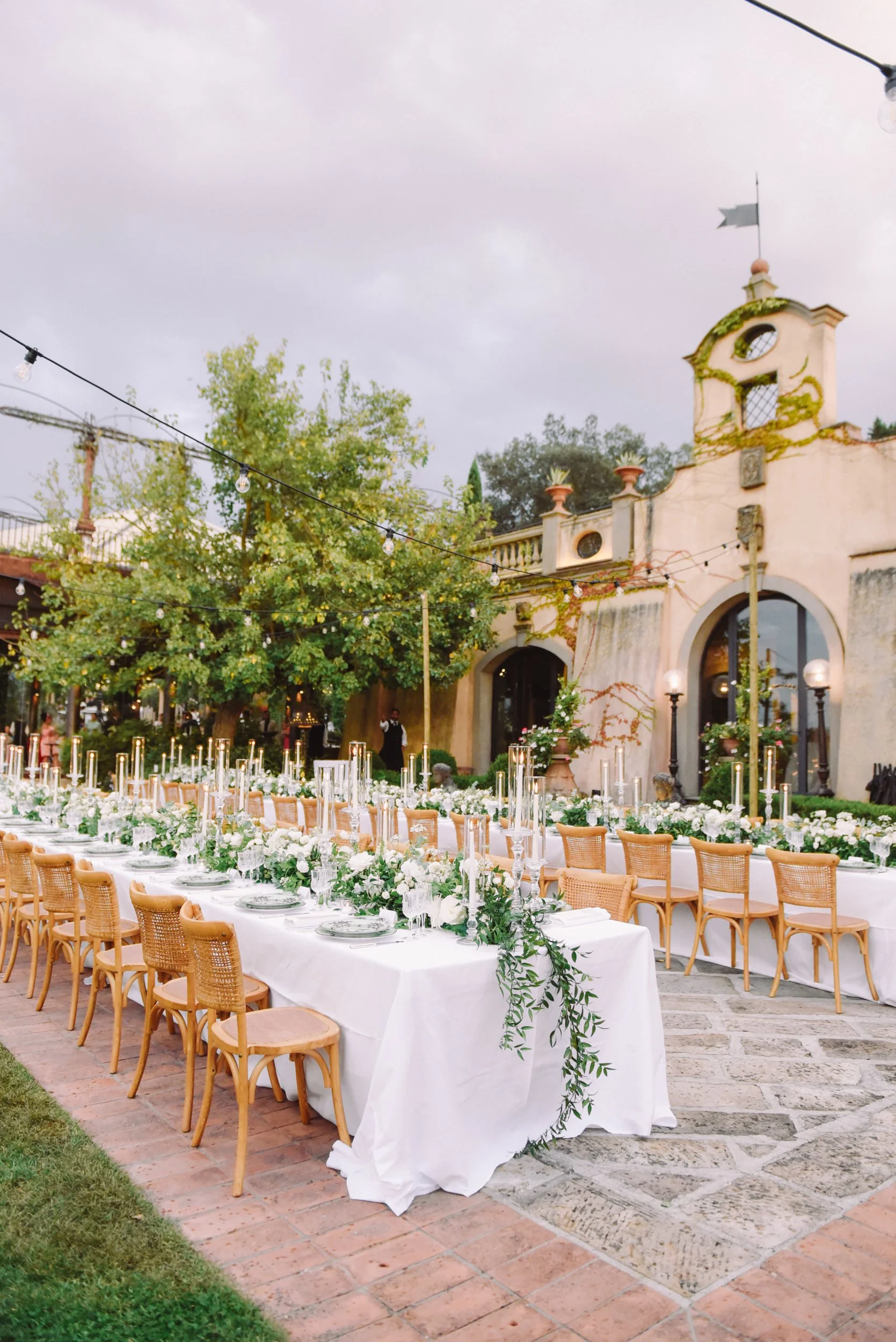 Outdoor wedding reception with long white tables, candles, and floral arrangements, set on a brick patio in front of an old building with ivy.