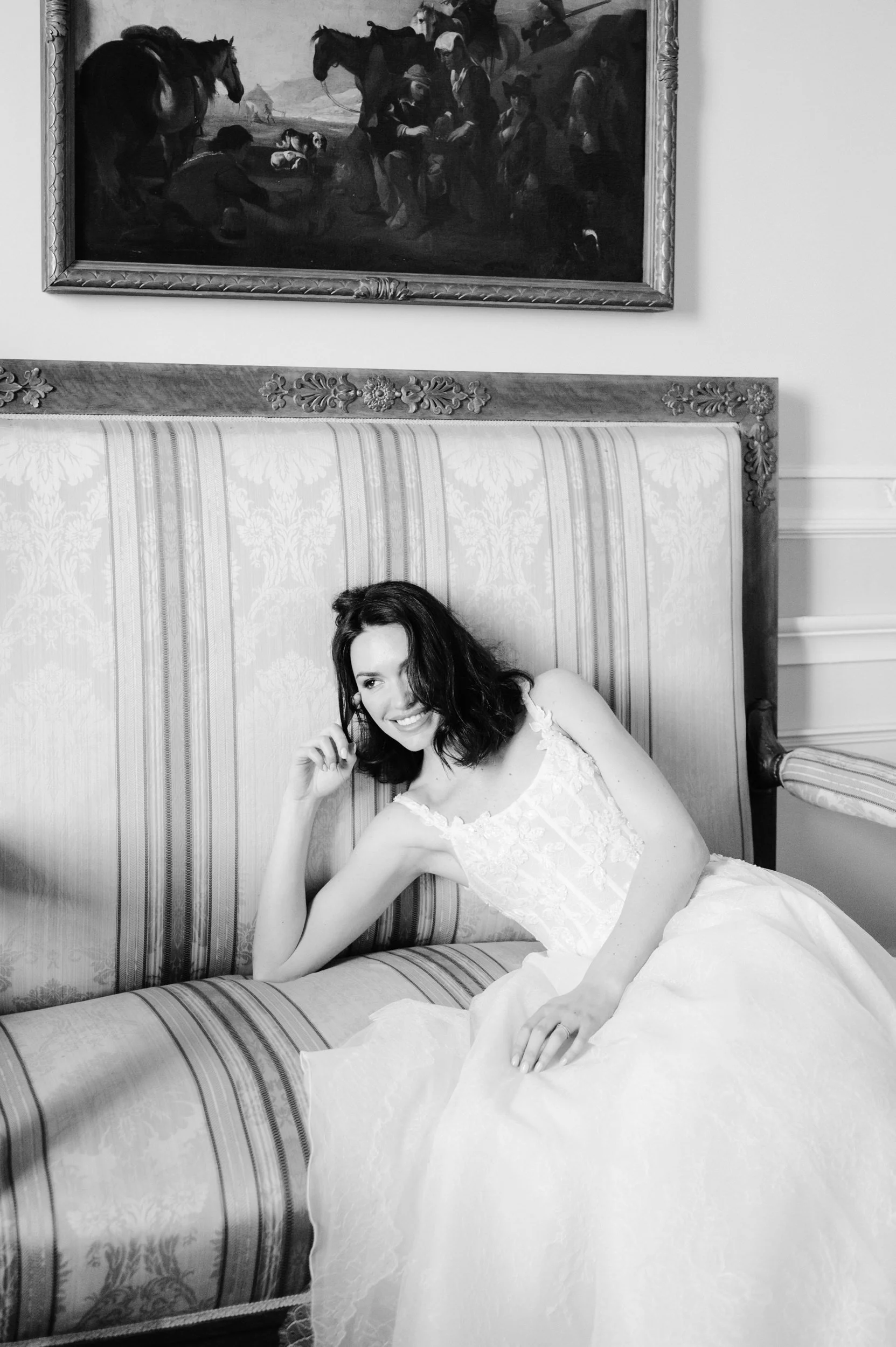 A woman in a white dress sitting on a vintage sofa, smiling and resting her head on her hand. There is a framed painting hanging on the wall above her.