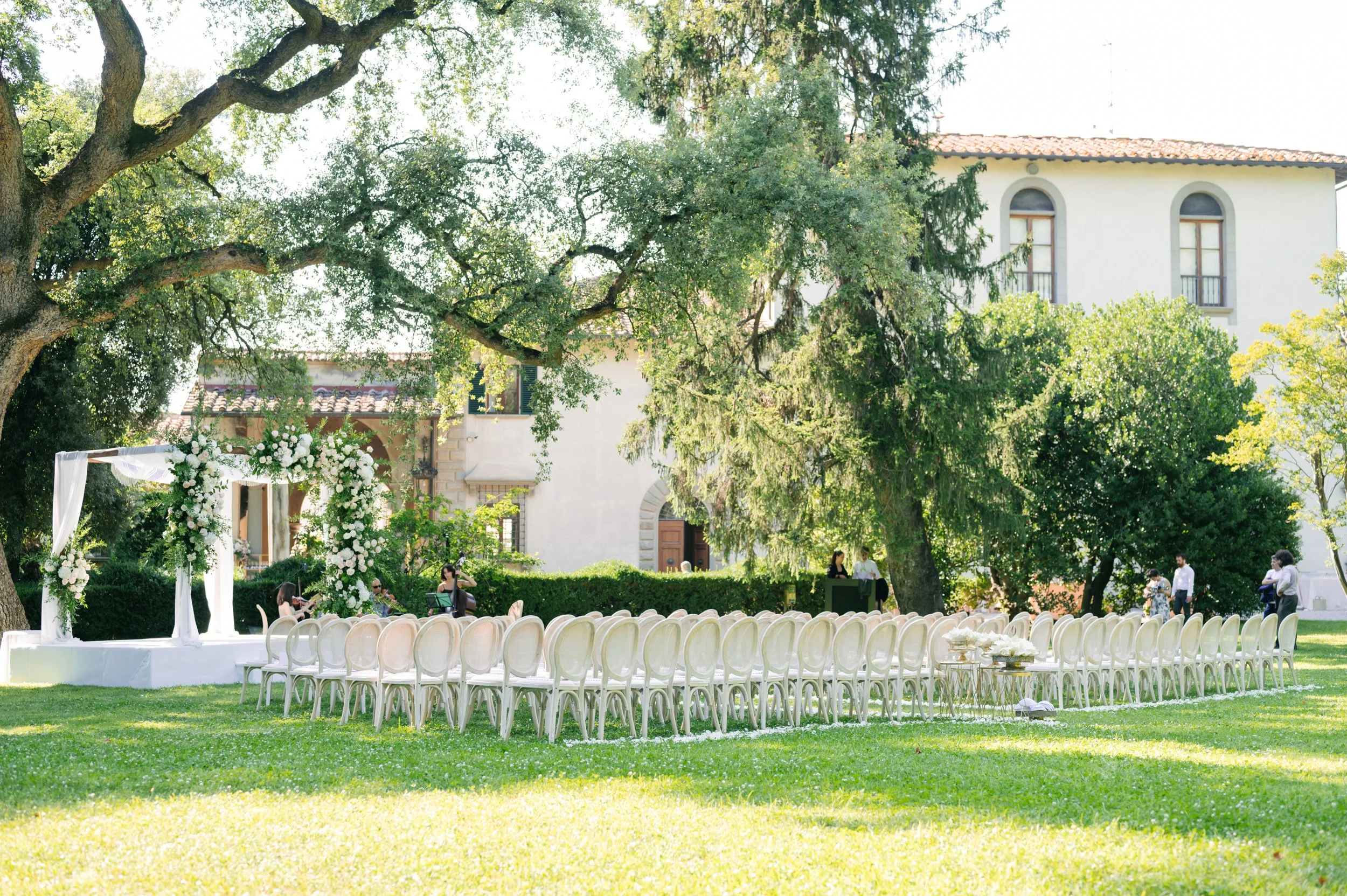 Outdoor wedding setup with a floral arch, white chairs arranged in rows on a green lawn, and a large tree providing shade, in front of a large white building with arched windows.