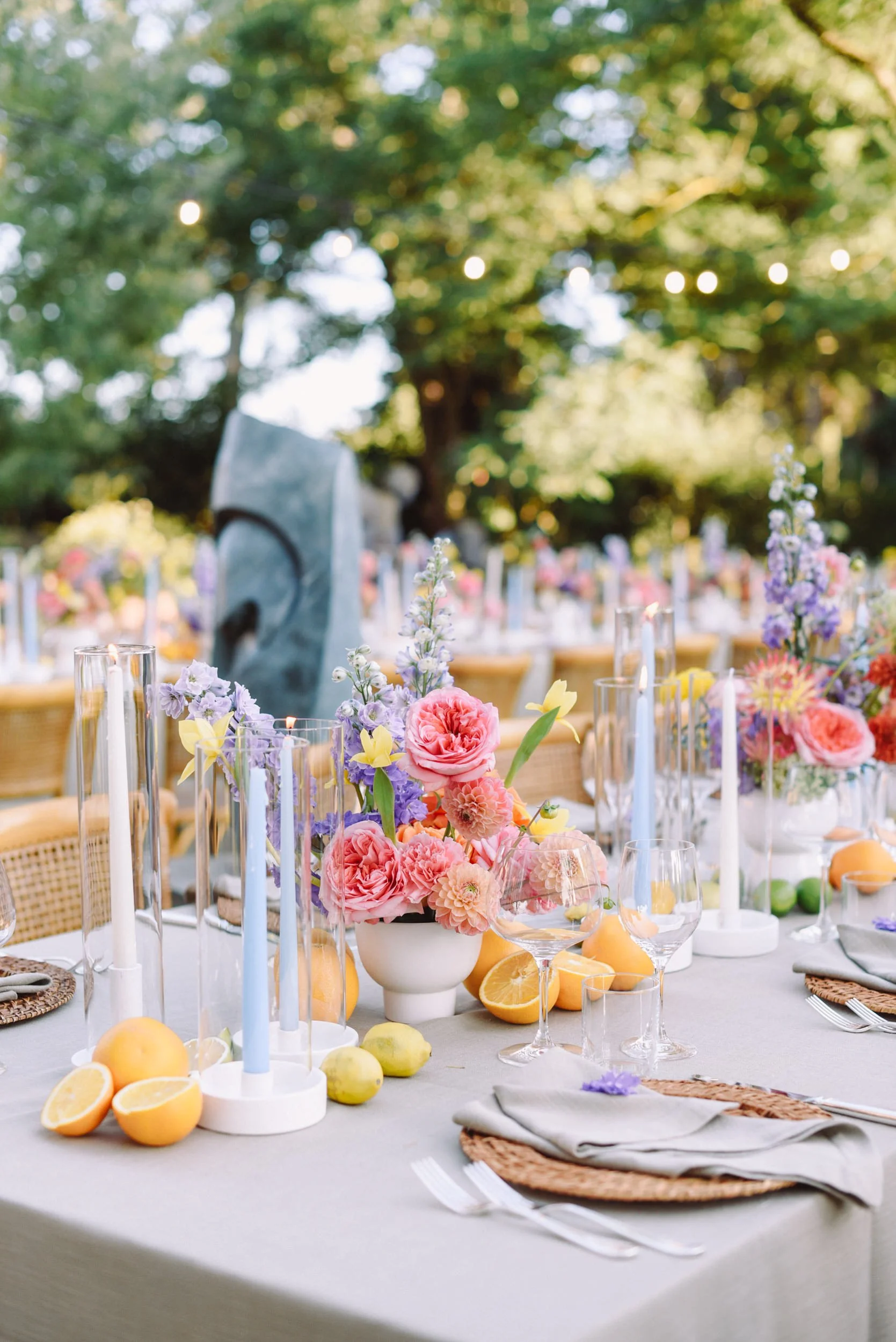 Outdoor event table decorated with pink, purple, and yellow flowers, surrounded by sliced citrus fruits and candles, with trees and string lights overhead.