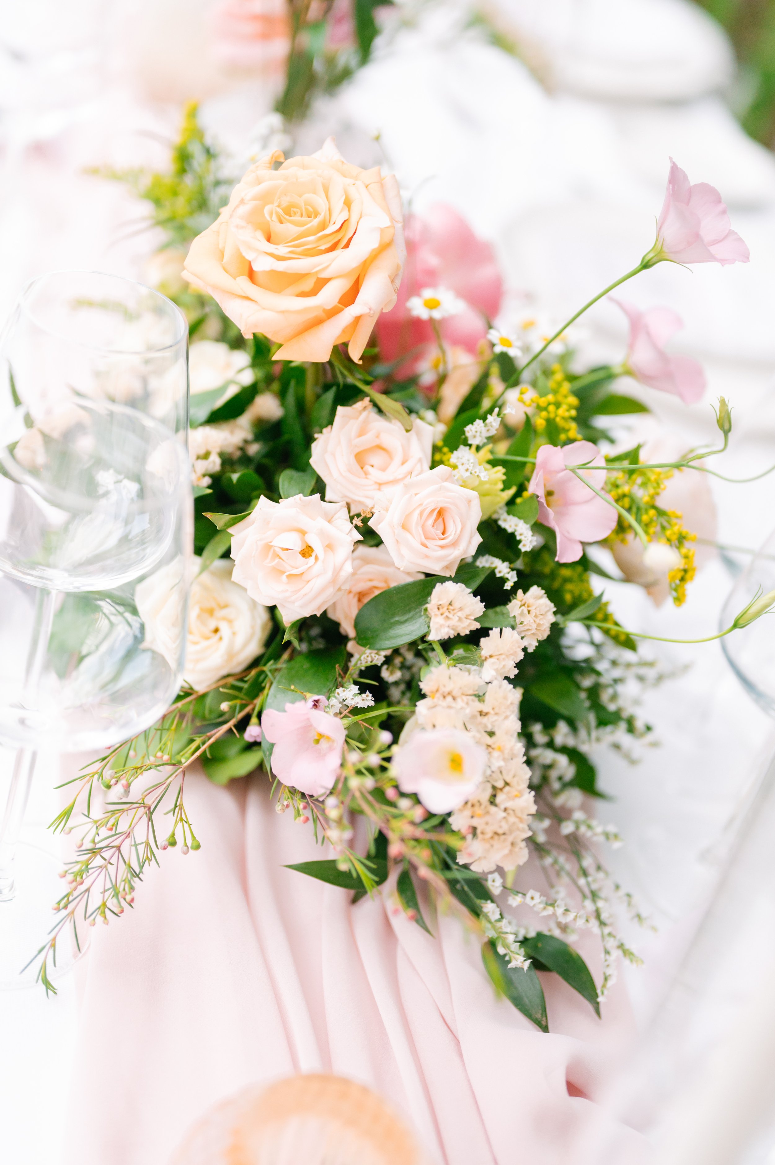 A floral centerpiece with peach roses, pink lisianthus, white daisies, yellow flowers, and greenery on a pink fabric tablecloth, with wine glasses nearby.