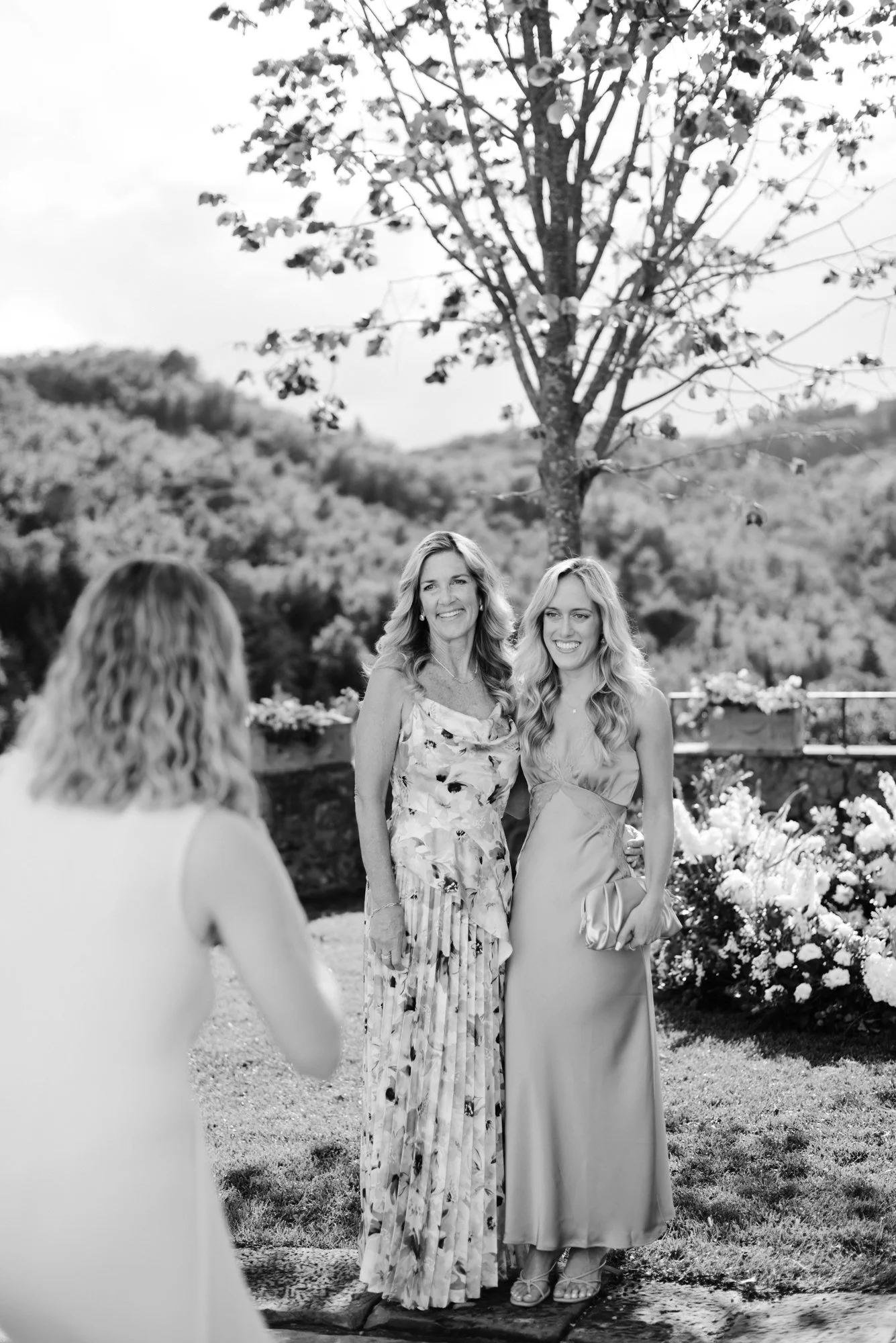 A photo of two women in formal dresses standing outdoors in front of flowering bushes and a tree, smiling for a picture. A third woman is taking the photo.