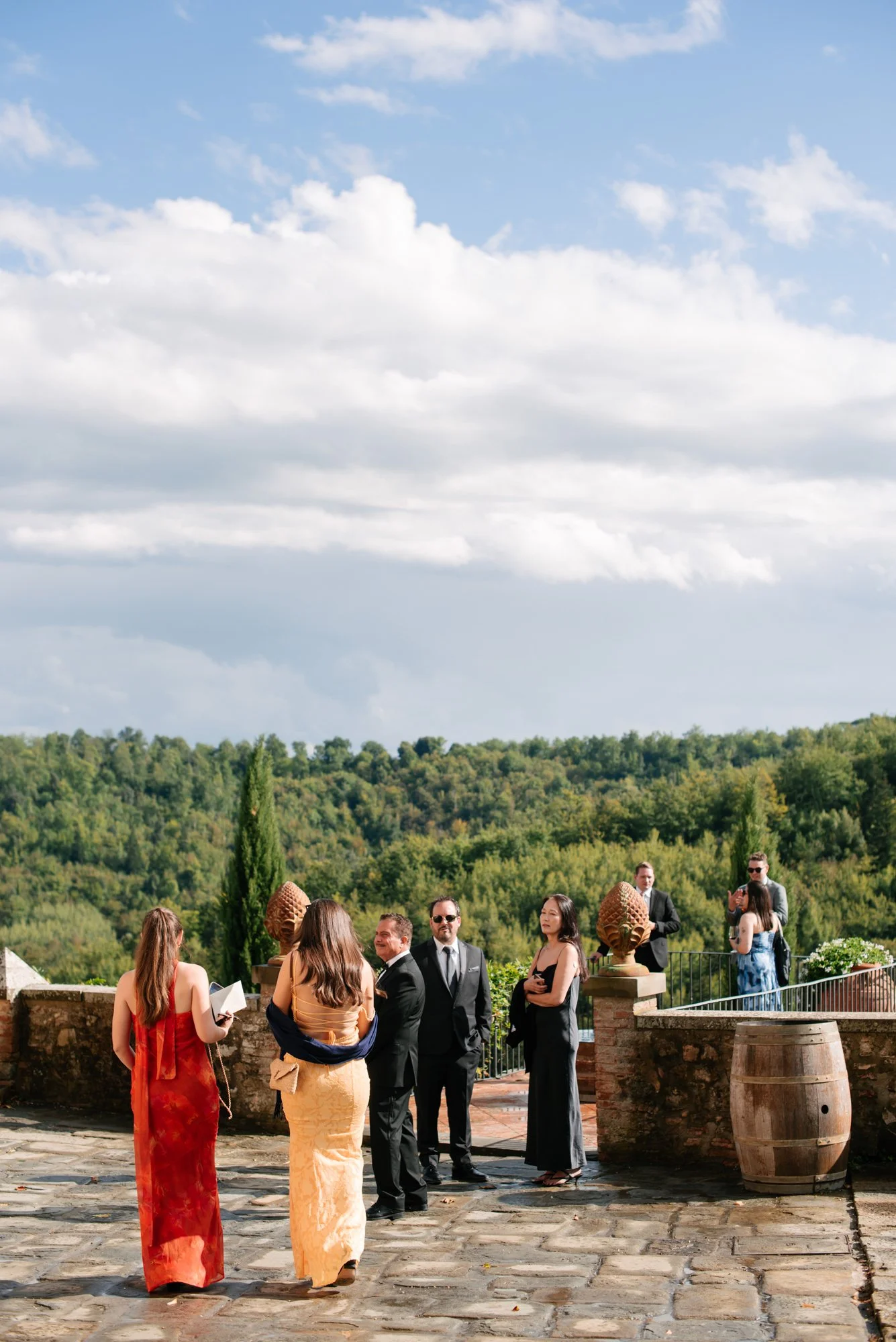 Group of people dressed in formal attire standing and conversing on a stone patio outdoors, with greenery and rolling hills in the background under a partly cloudy sky.
