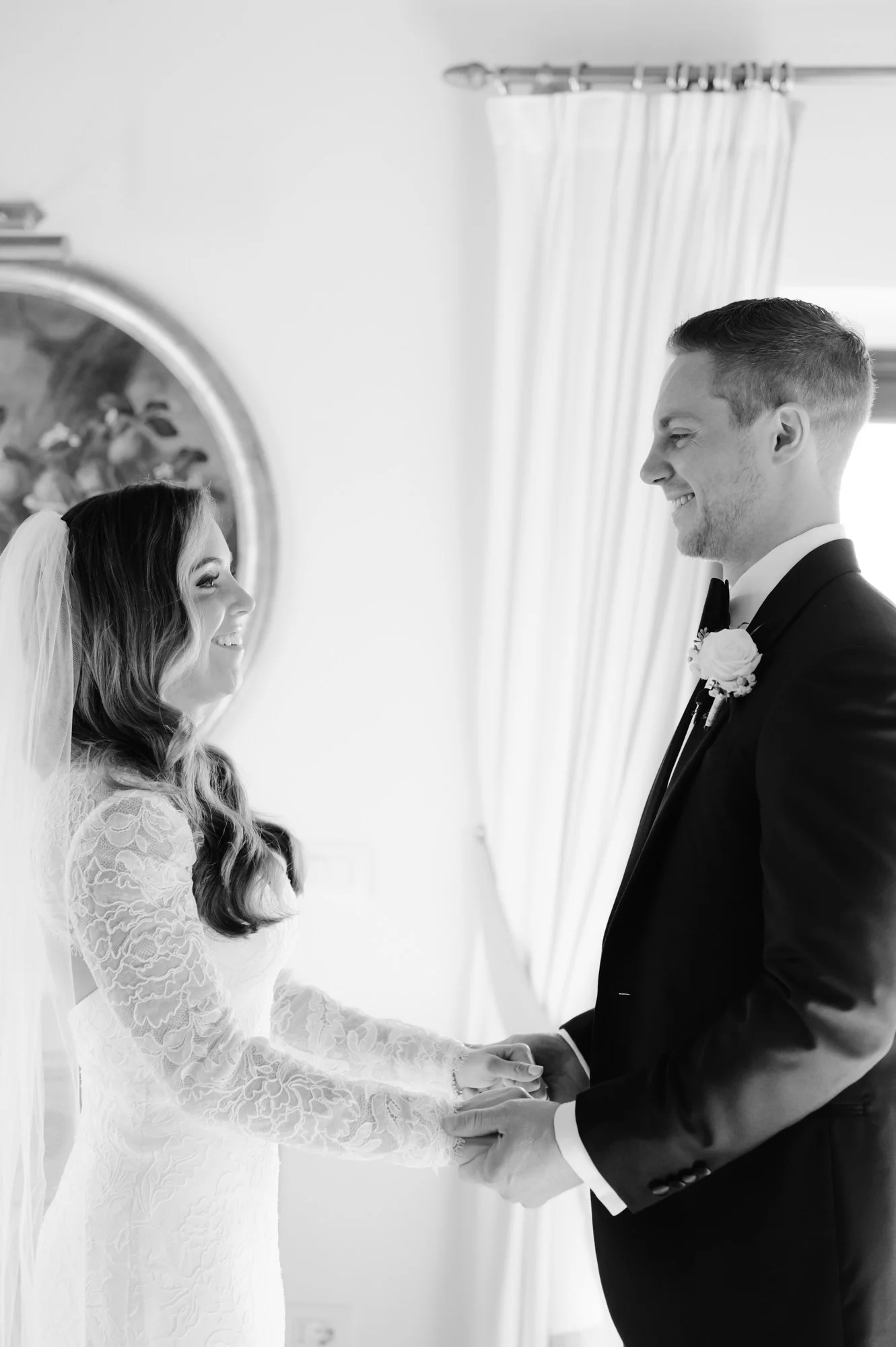 A bride and groom holding hands and smiling at each other during their wedding ceremony inside a room with curtains and a decorative mirror.