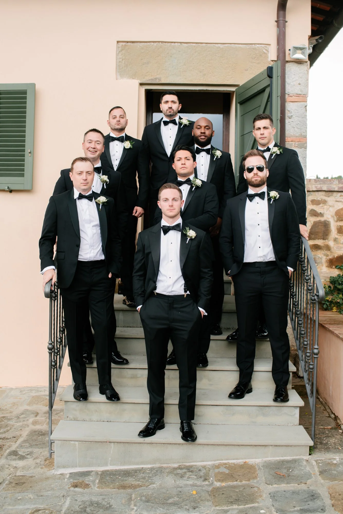 A group of men dressed in black tuxedos with bow ties, standing on a staircase outside a building, some with floral boutonnieres. Luxury editorial wedding photography in Tuscany. Destination wedding in Italy. L&V Photography Letizia Maccarini.