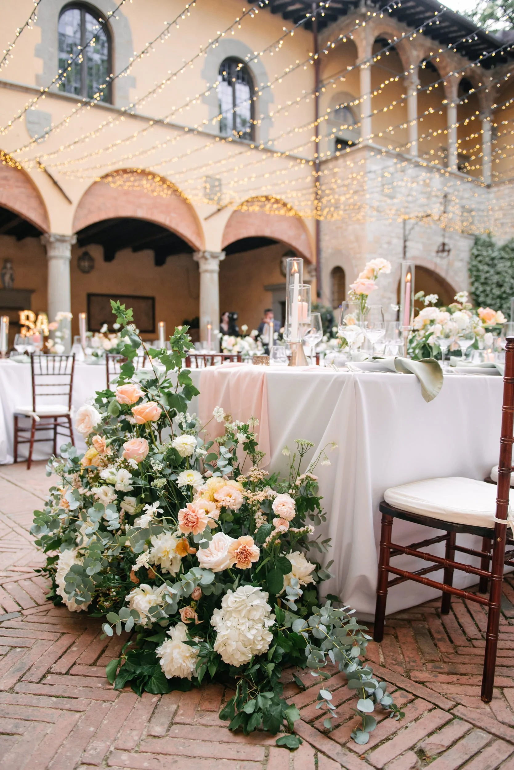 Elegant outdoor wedding reception with long tables decorated with white tablecloths, floral arrangements, and tall candle holders. Overhead string lights and a historic stone building with arched windows and columns create a romantic ambiance.