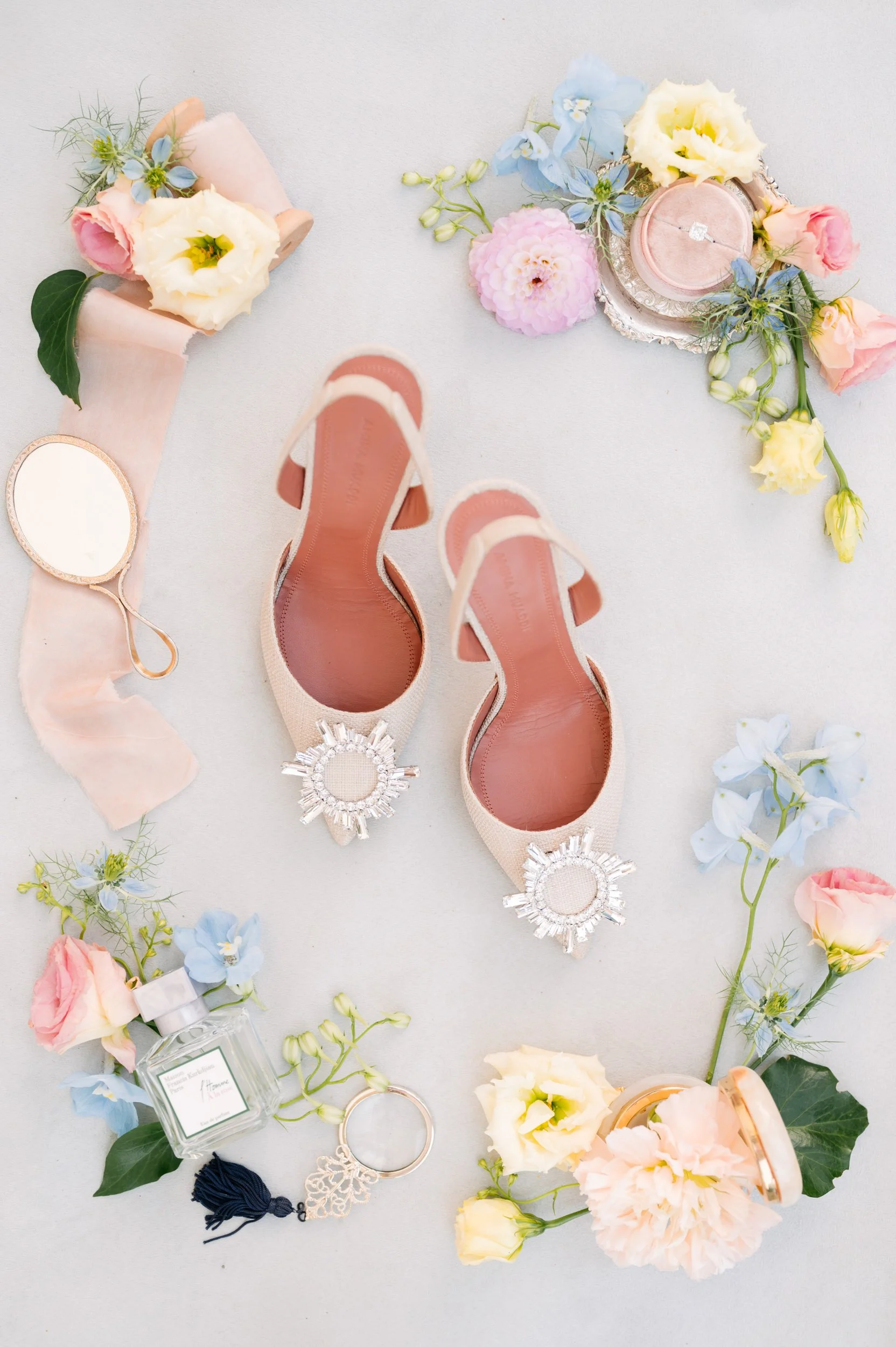 A flat lay of wedding accessories including a pair of blush pink heels with jeweled embellishments, pink and yellow flowers, a small mirror, a perfume bottle, a ring, and a candle, arranged on a light background.