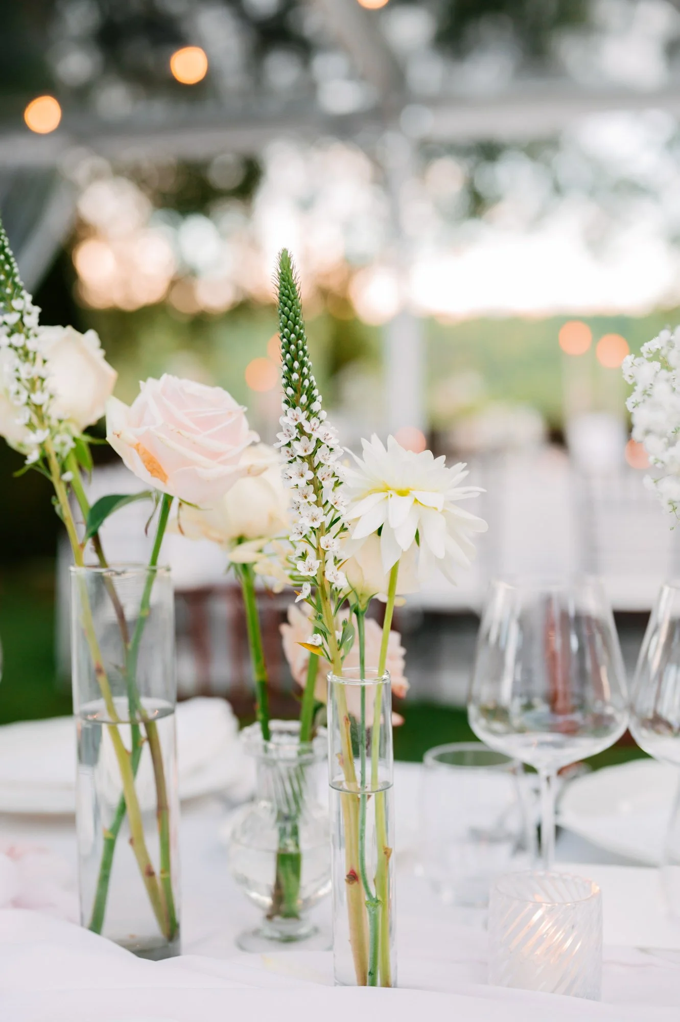 Elegant floral centerpiece with white roses, dahlias, and tall white delphinium in clear glass vases on a table at an outdoor event