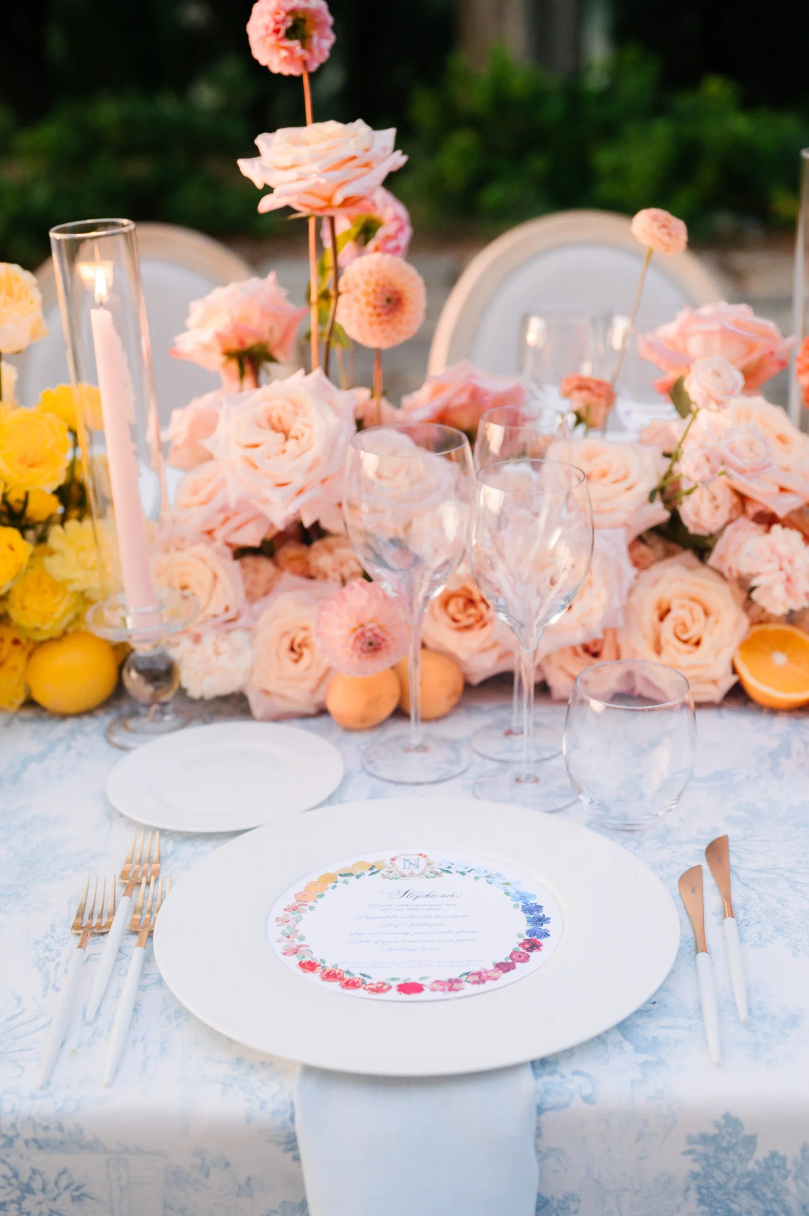 Elegant outdoor table setting decorated with pink roses, peach and yellow flowers, and glassware, with a menu on a white plate.
