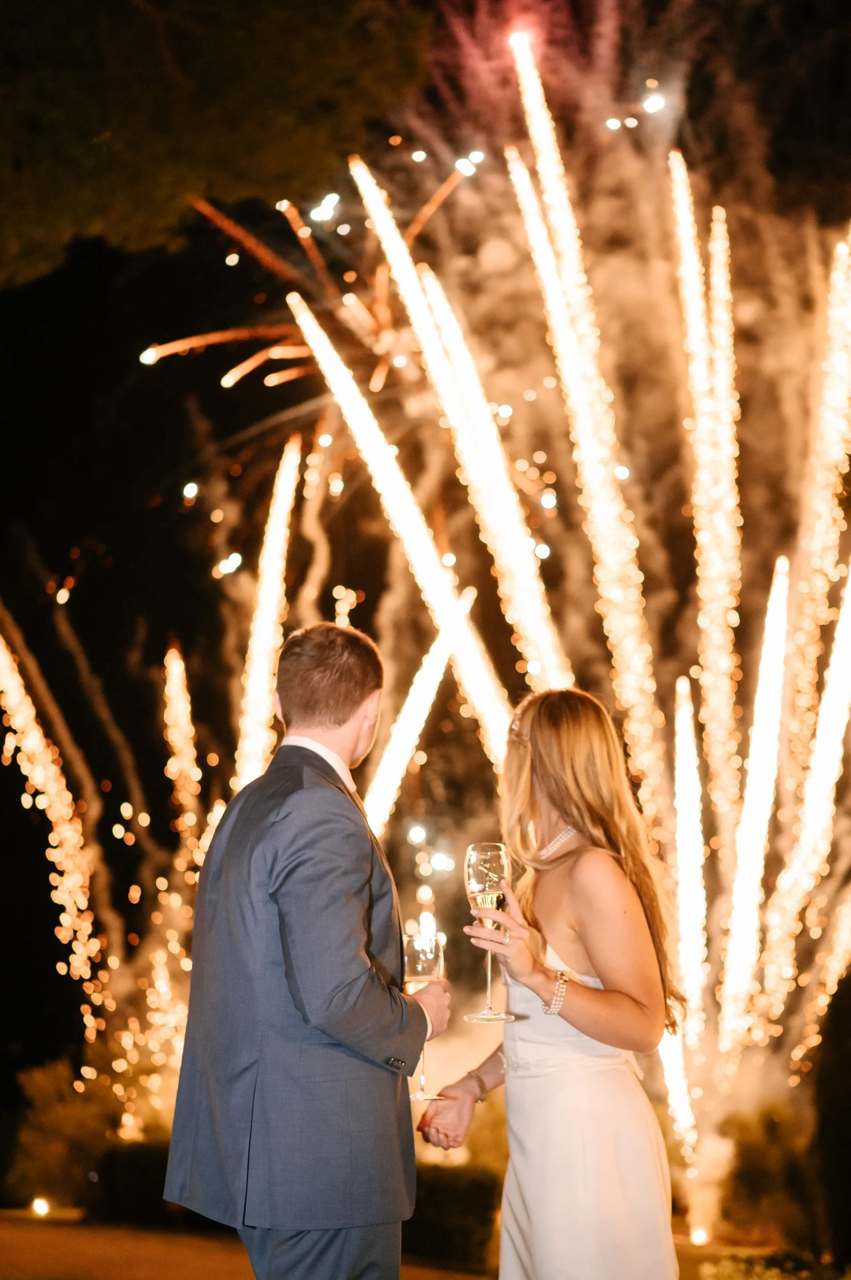 A couple in wedding attire holding glasses of champagne and holding hands during a fireworks celebration at night.