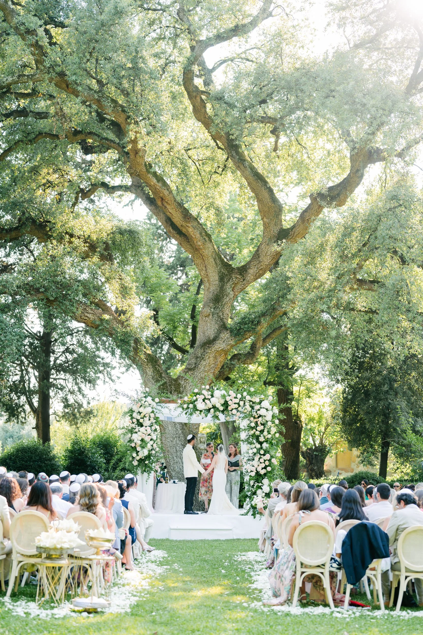 Outdoor wedding ceremony under a large tree decorated with white flowers, with guests seated on both sides.